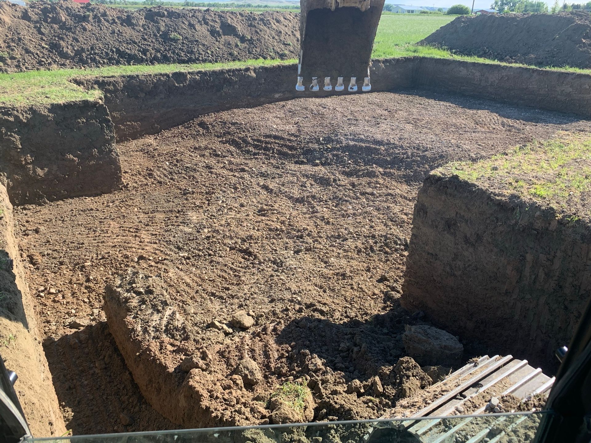 An excavator bucket is suspended over a deep rectangular pit excavated in an open, grassy field.