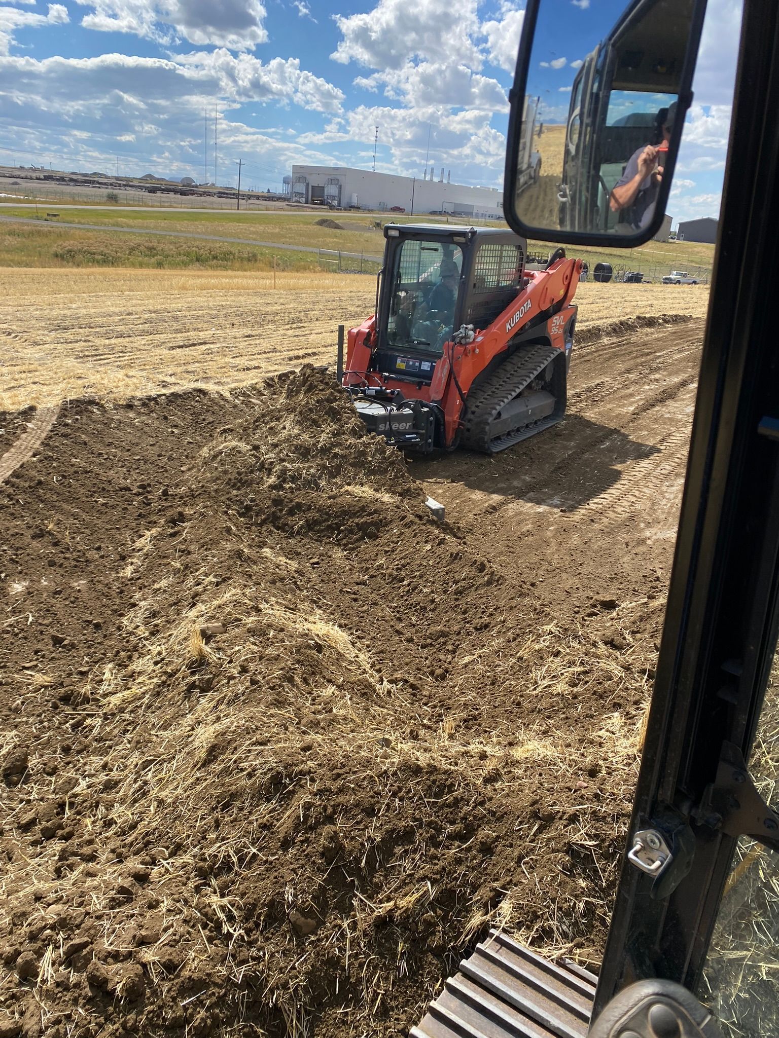 An orange skid steer loader operates in a field of tilled soil under a cloudy blue sky.