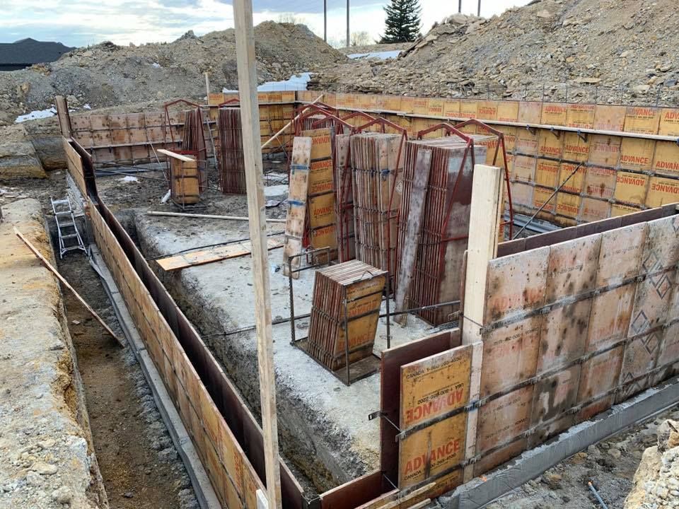 A yellow front-loader lifts a large black septic tank with orange straps while a worker guides it into an excavated trench.