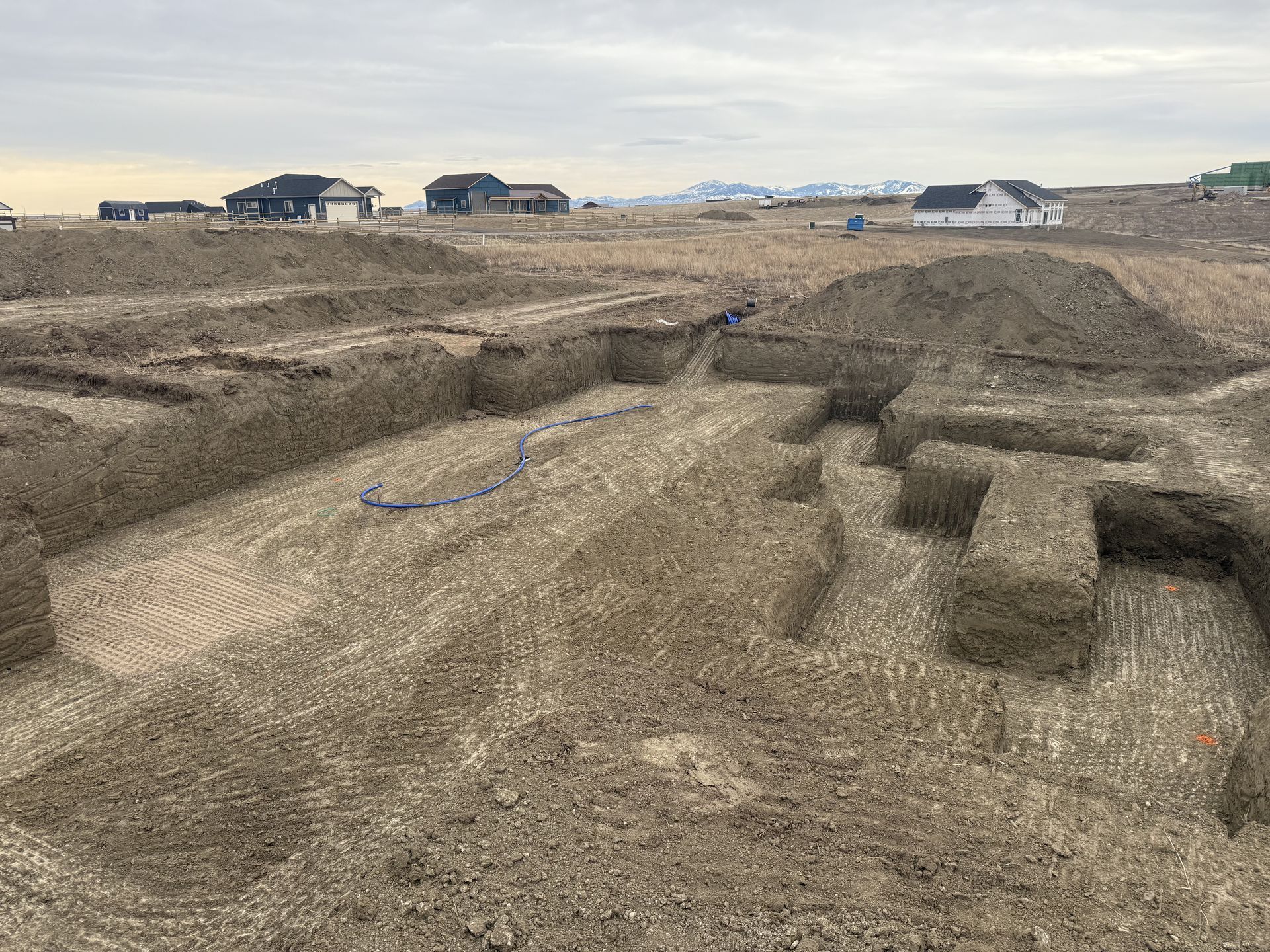 An excavated construction site showing deep, angular trenches carved into dry, brown earth with houses in the distance.