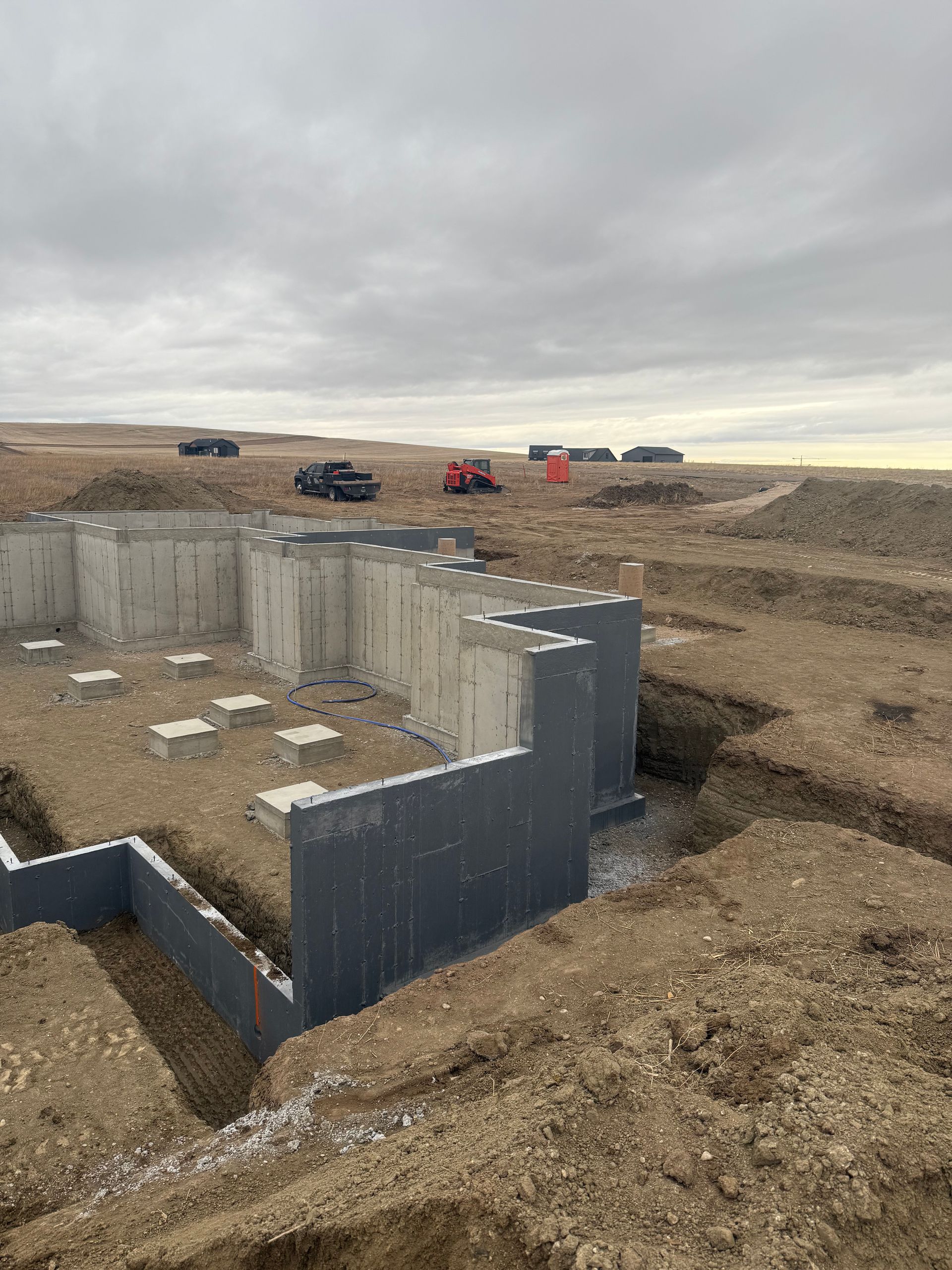 Concrete foundation walls for a building under construction on a dirt lot under a cloudy sky.