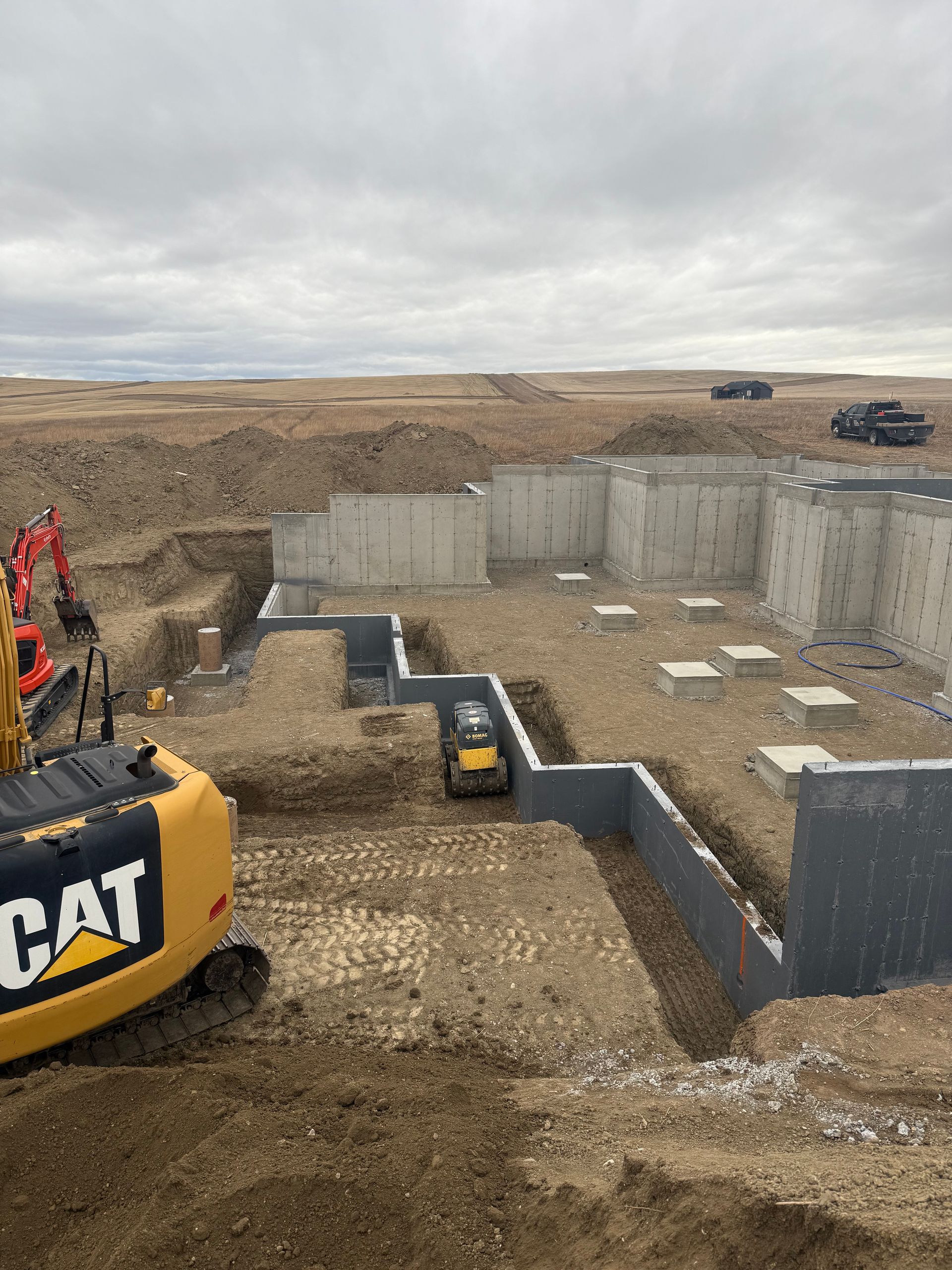 A construction site shows a concrete foundation wall with footings in a large, excavated dirt lot under a cloudy sky.