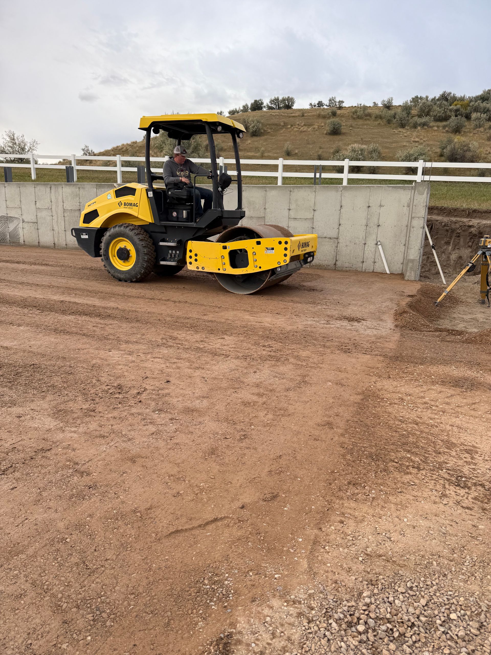 A yellow road roller compacting dirt in front of a concrete retaining wall with a white fence on a hillside.