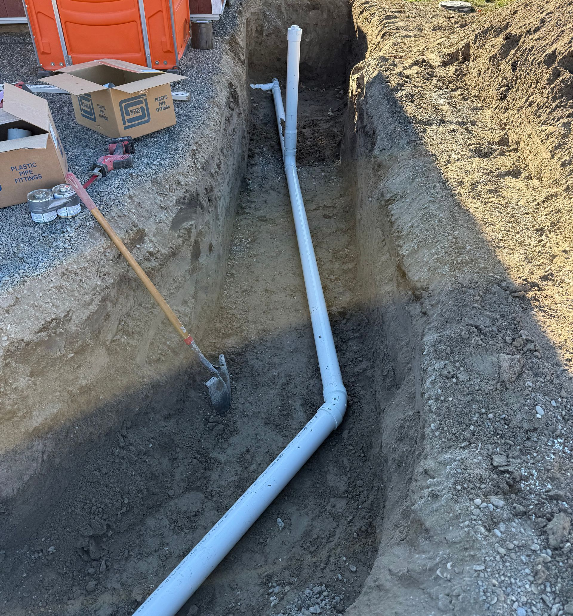 An excavator works in the background of a site featuring two rows of black, corrugated plastic drainage pipes in trenches.
