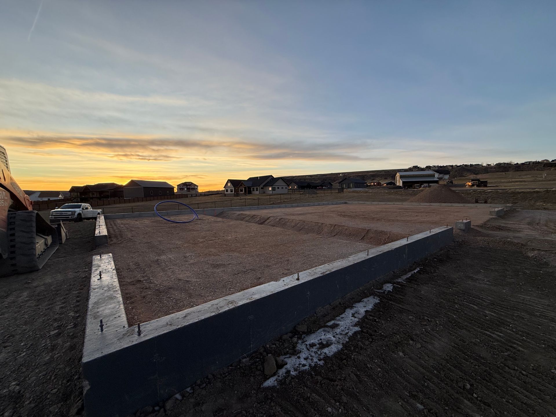 A concrete foundation for a new house at a construction site during a sunset with other houses in the distance.