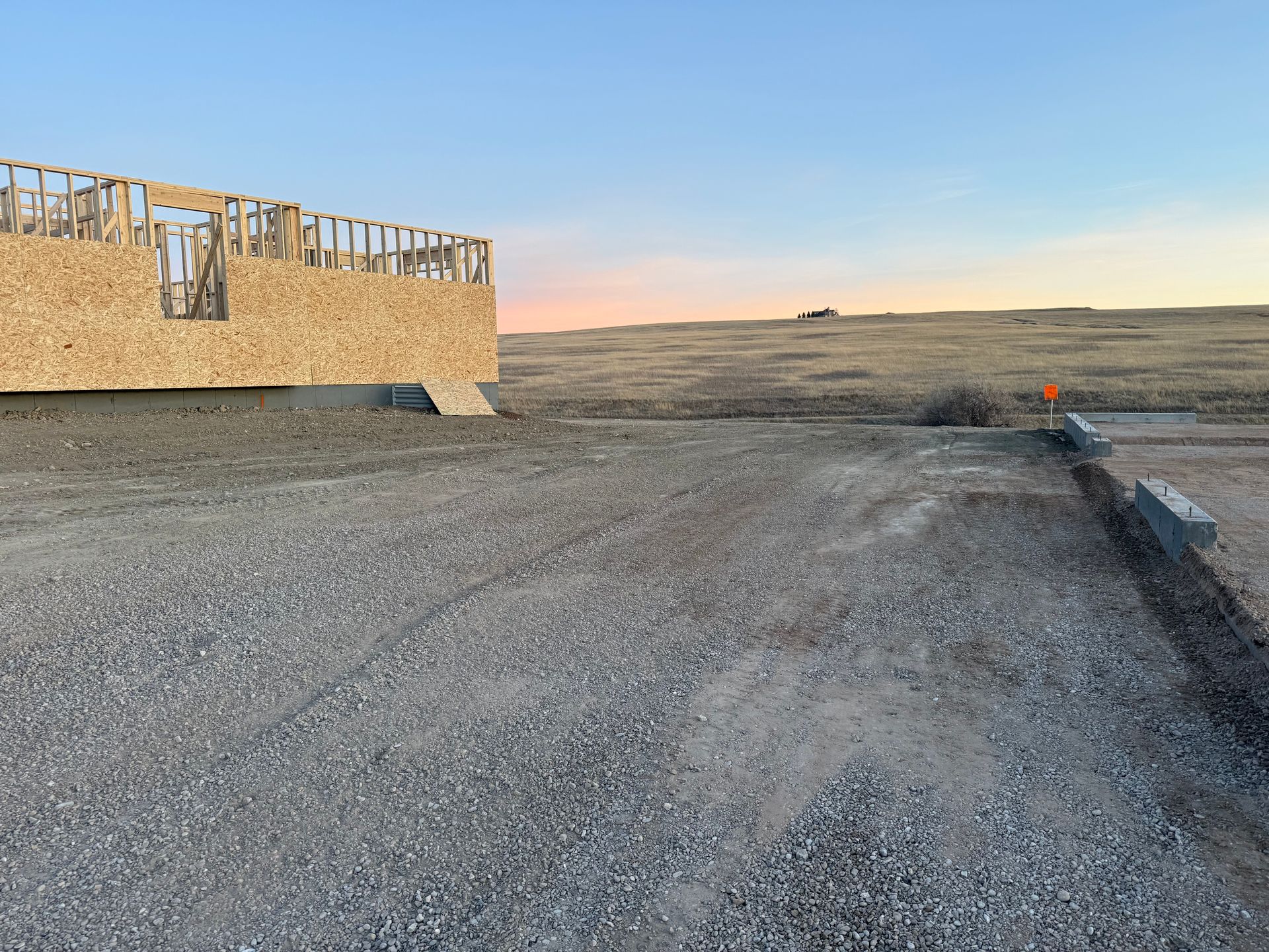 Construction site with a partially framed wooden building on a gravel lot under a pale sunset sky.