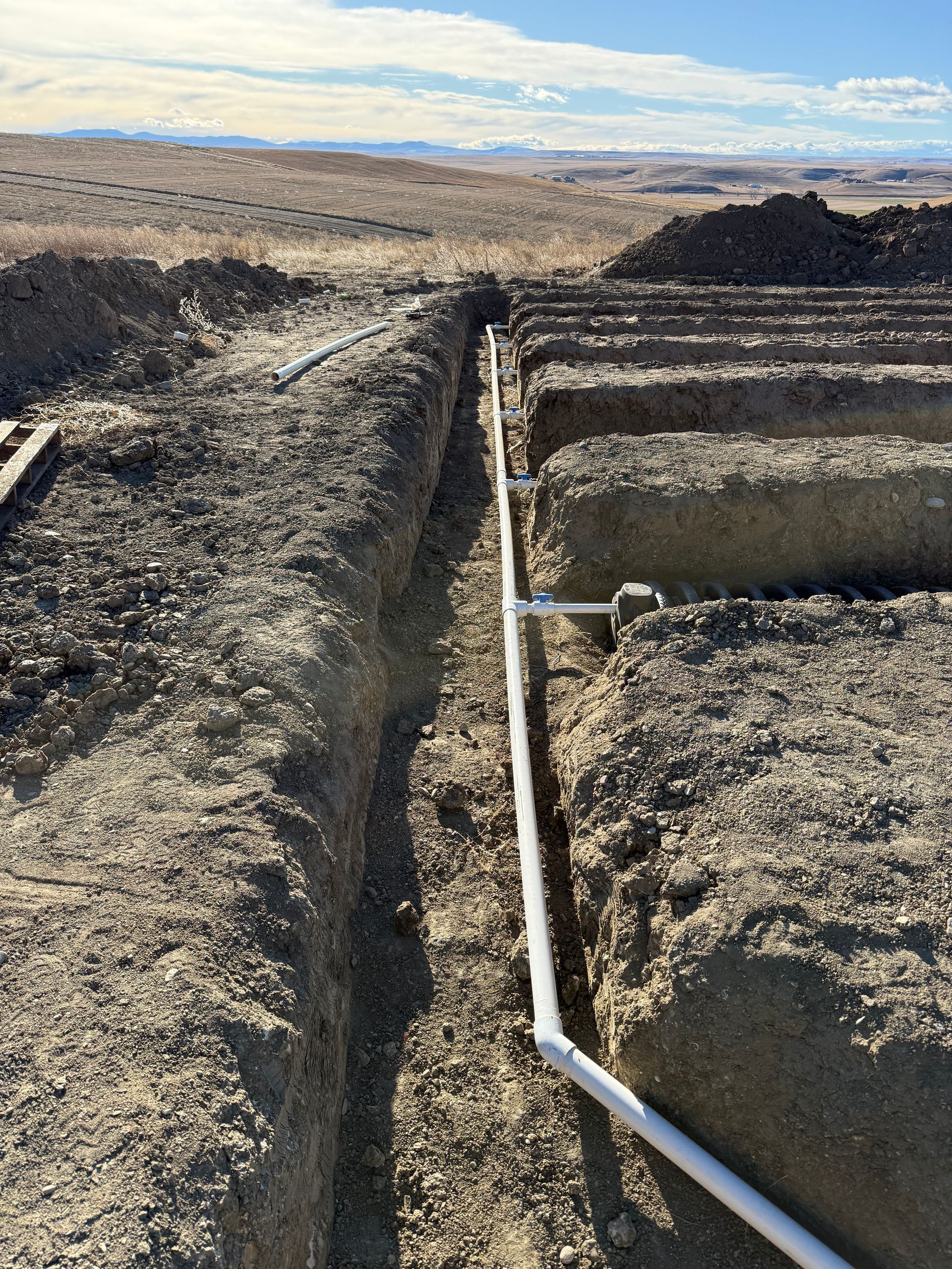 A white PVC pipe runs along a trench beside parallel earthen mounds in a desert landscape under a blue sky.