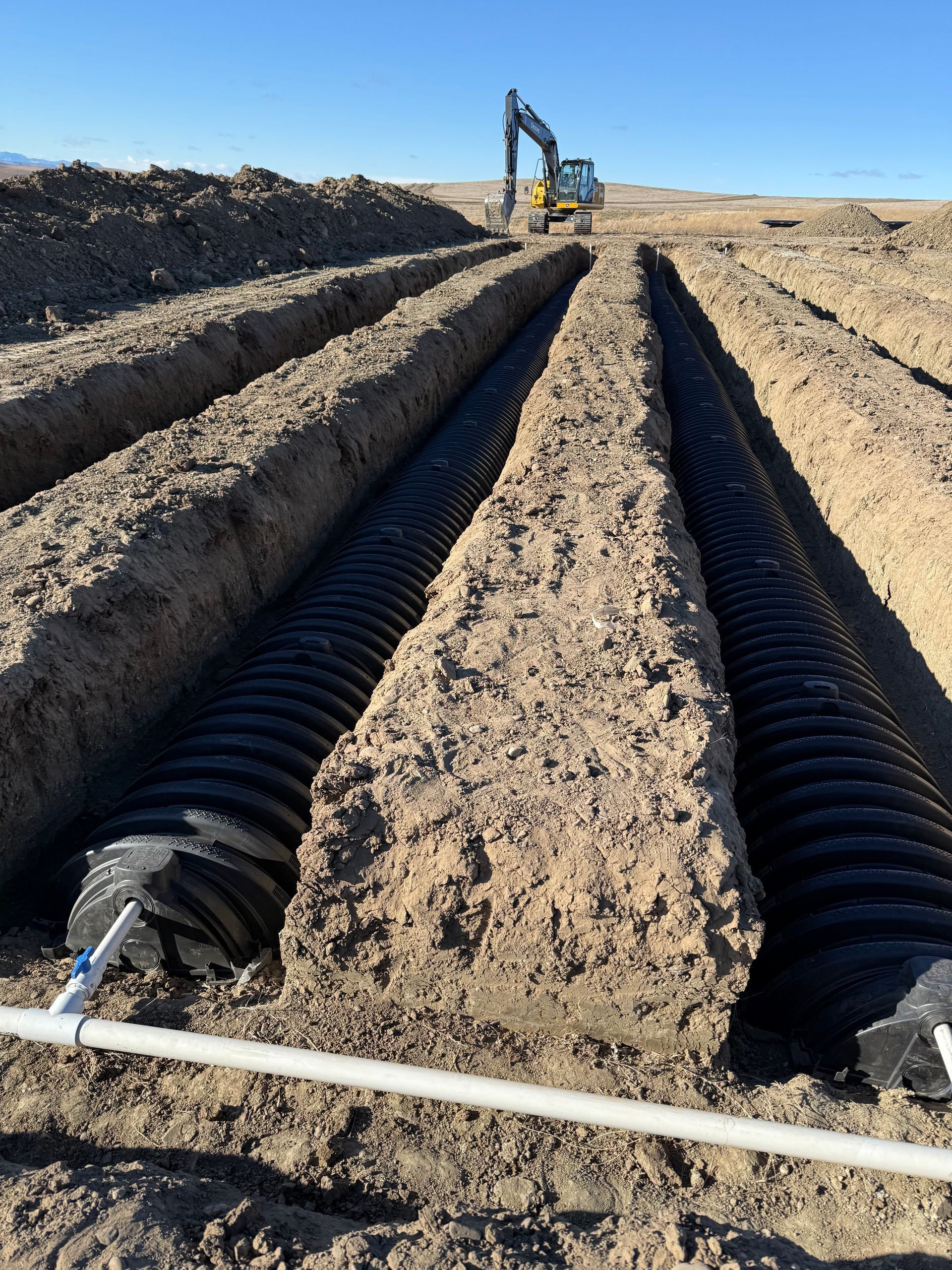 An excavator works near two parallel rows of black plastic septic chambers installed in trenches in a dirt field.