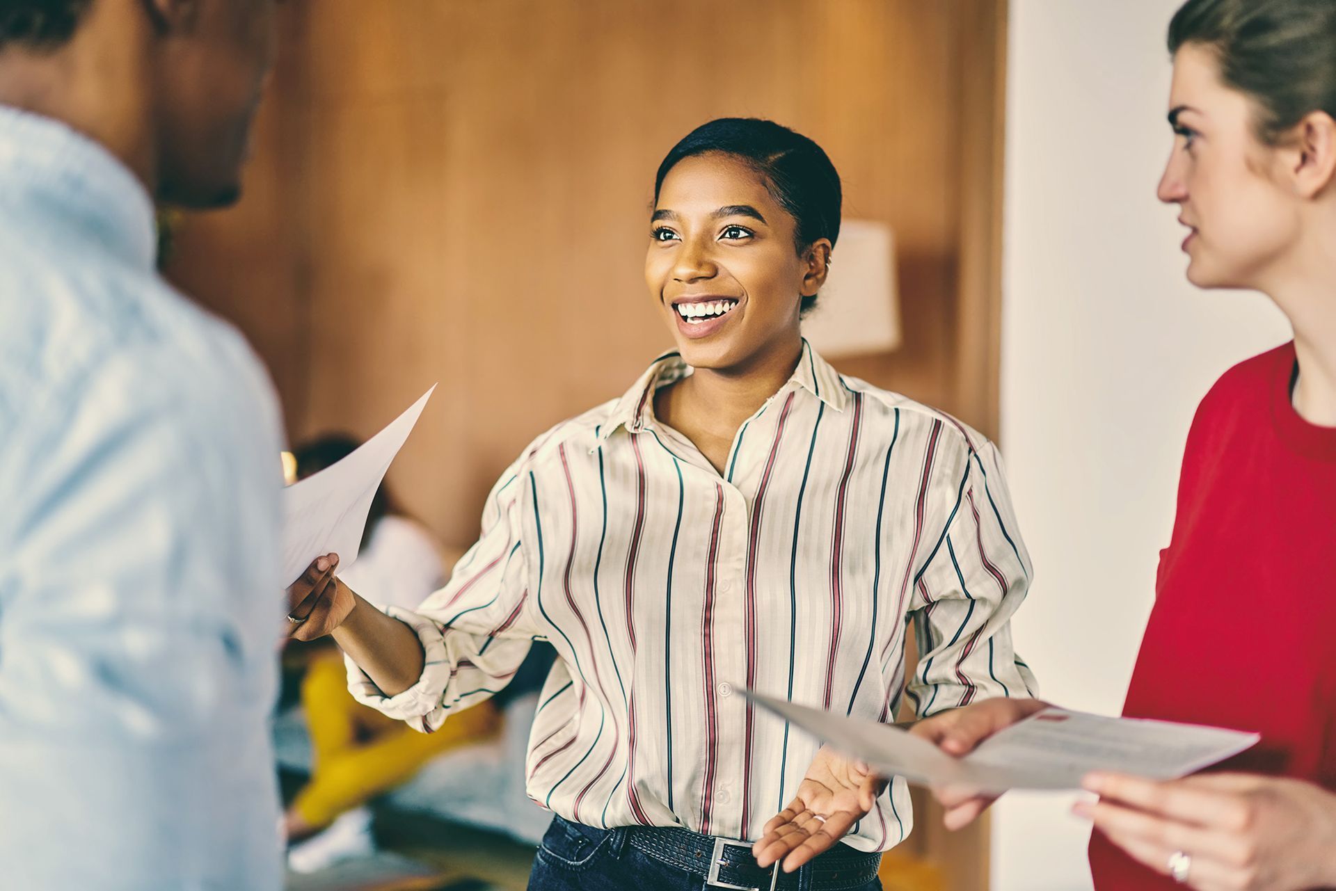Woman smiles, holding papers, talking to two colleagues in an office setting.