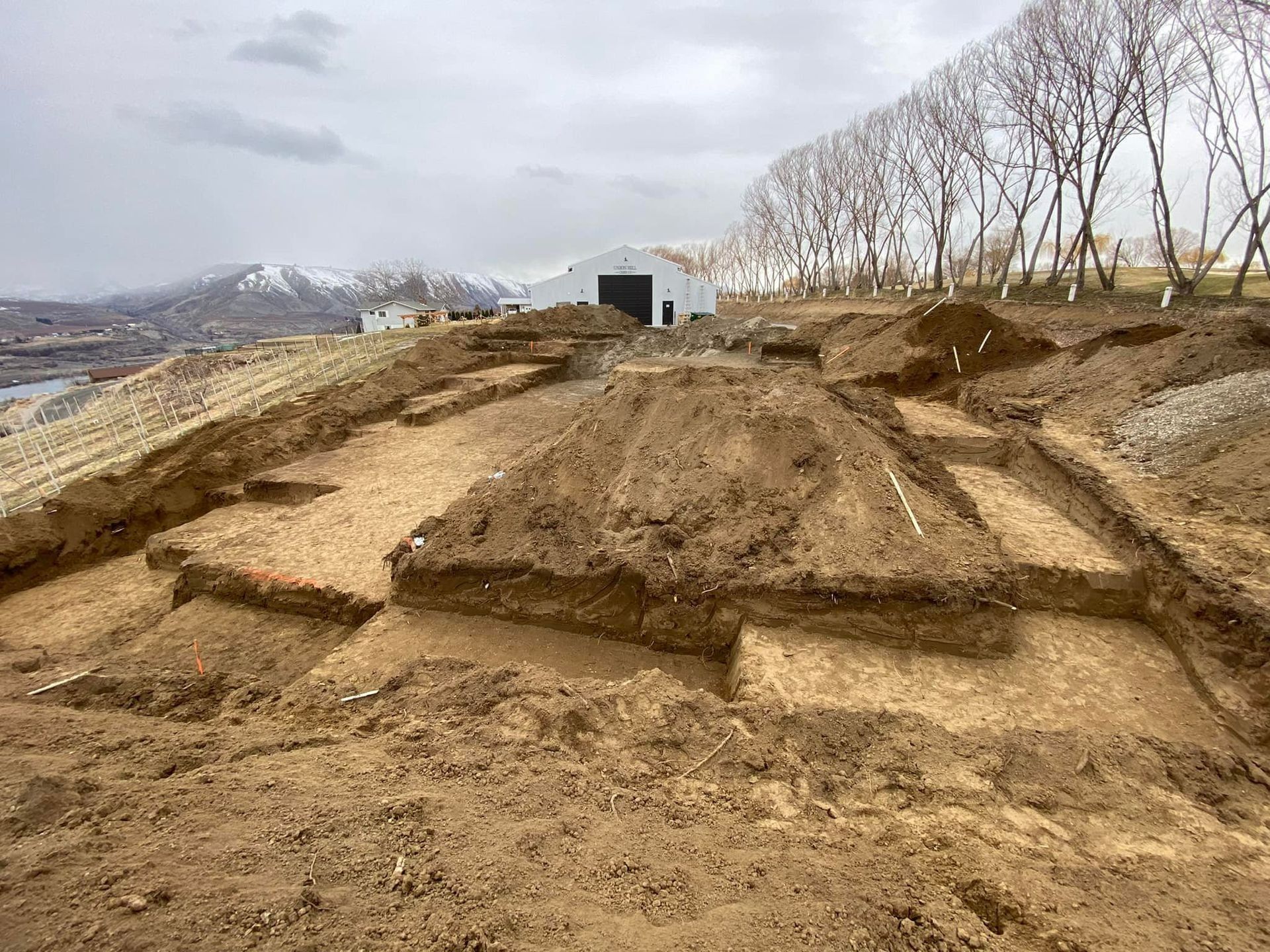 Excavation site with rectangular trenches in brown soil. In the background, mountains and a building are visible.