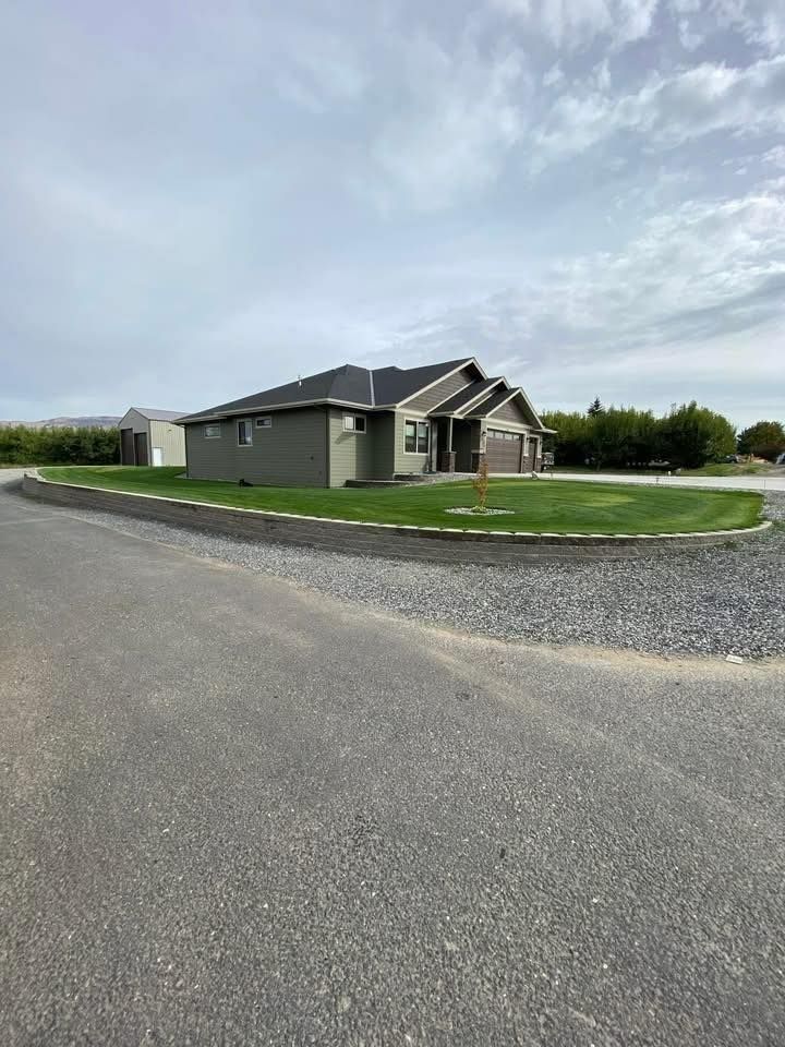 Single-story house with green siding and dark roof, set on a gravel driveway, under a cloudy sky.