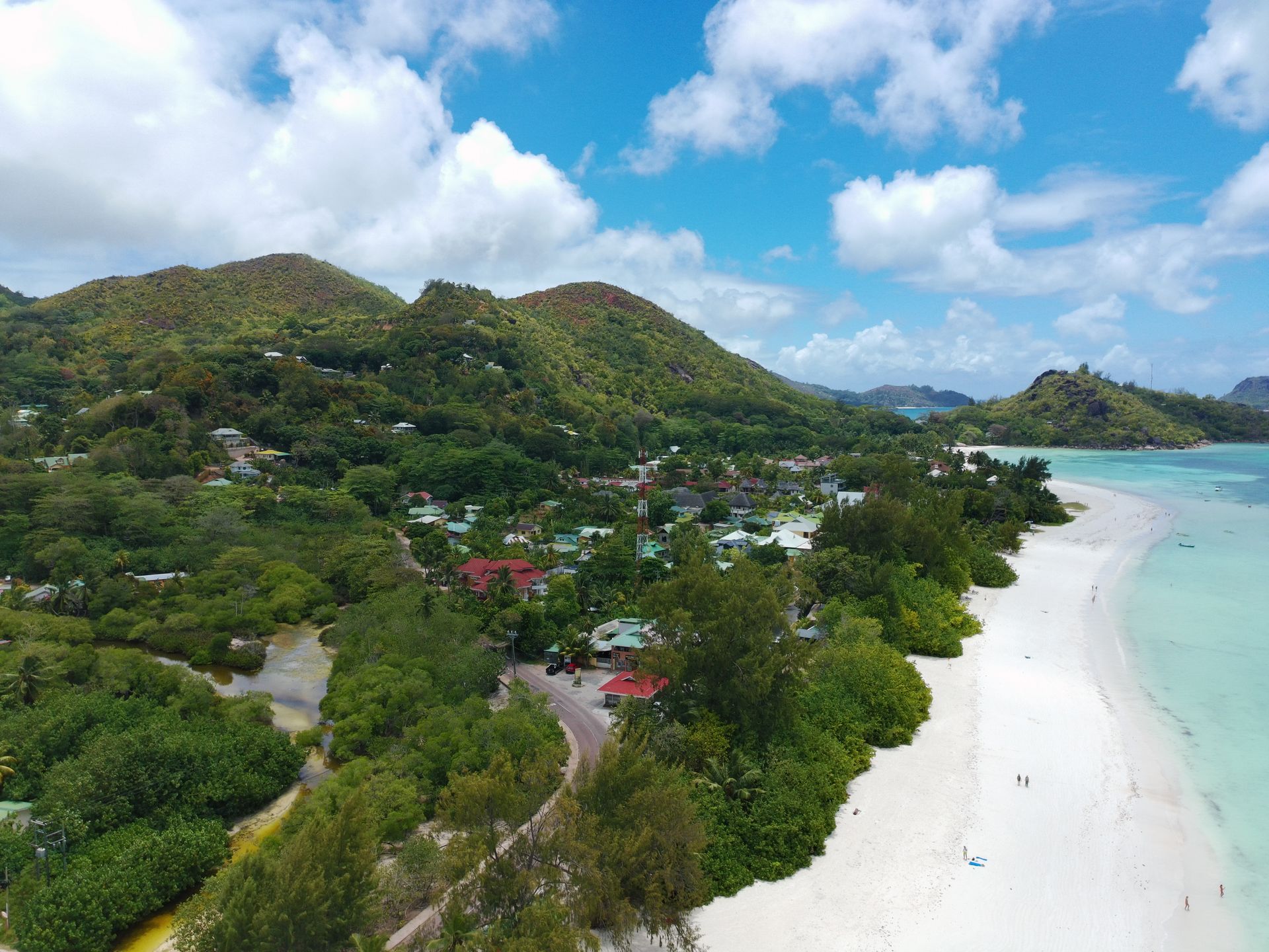 White sand beach, turquoise water, lush green hills, and a village under a blue sky with clouds.