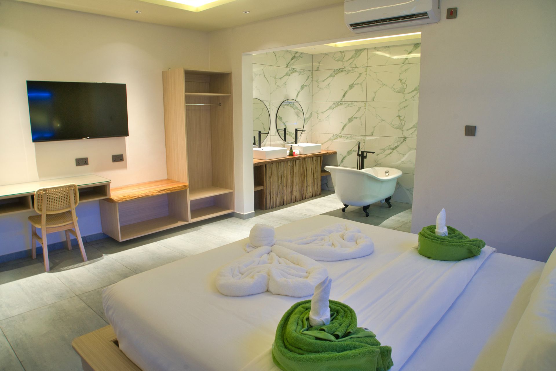 Hotel room with bed, TV, desk, closet, and a bathroom with a tub. White walls, wood accents.