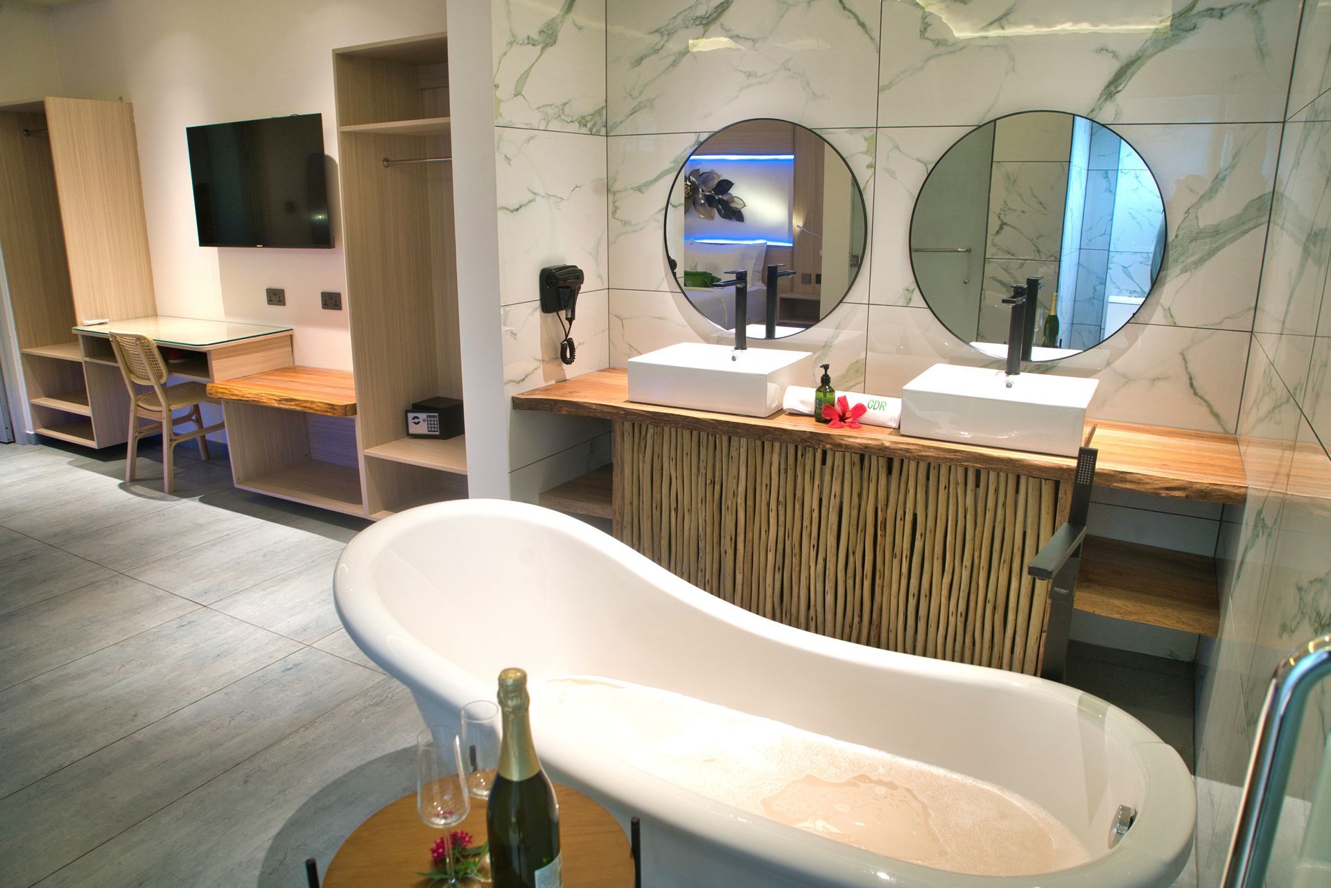 Bathroom with a freestanding tub, dual sinks, and a desk area with a TV, featuring light wood and marble accents.