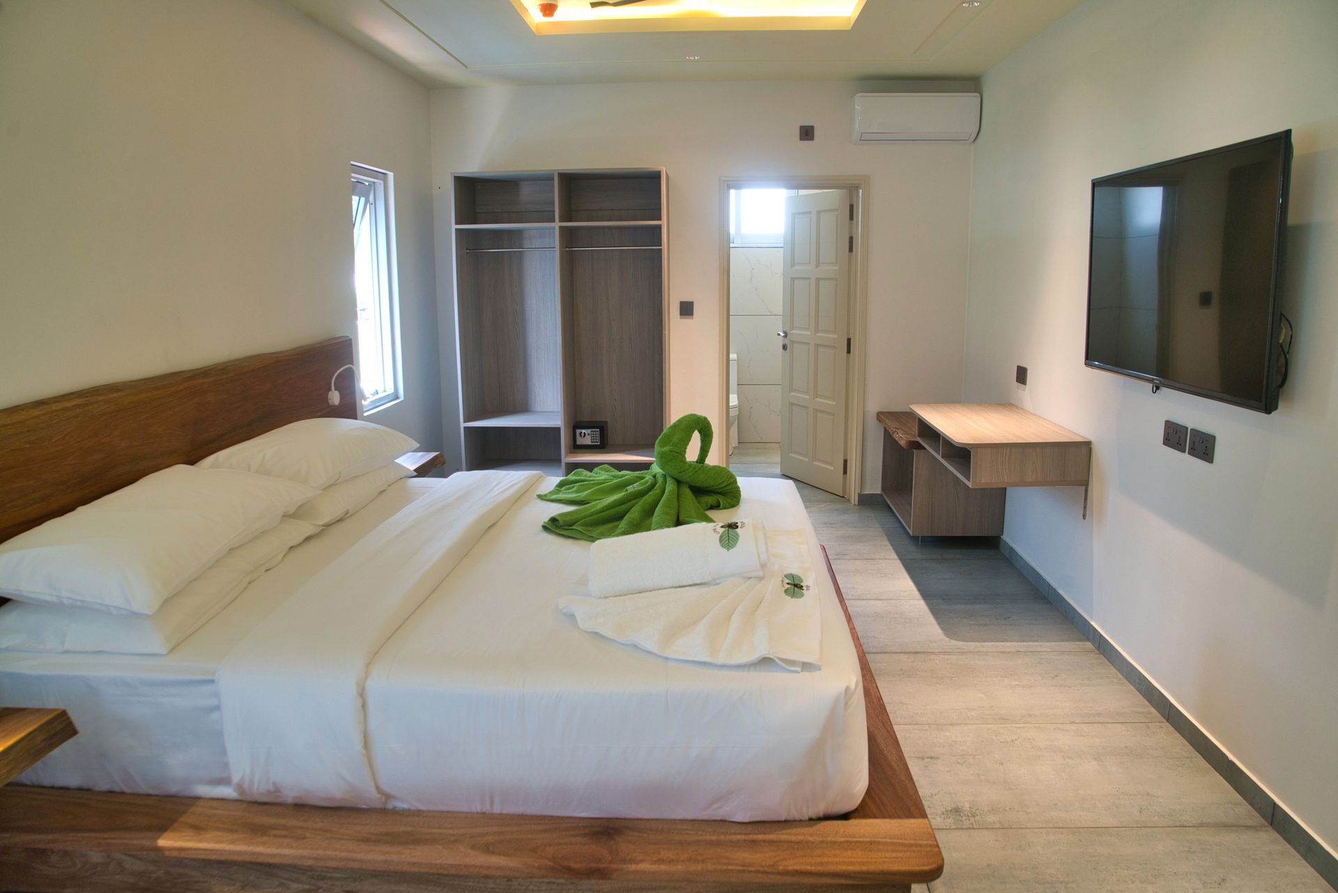 Bedroom with bed, closet, TV, desk, and open door to bathroom. White and wood tones with green towel decorations.
