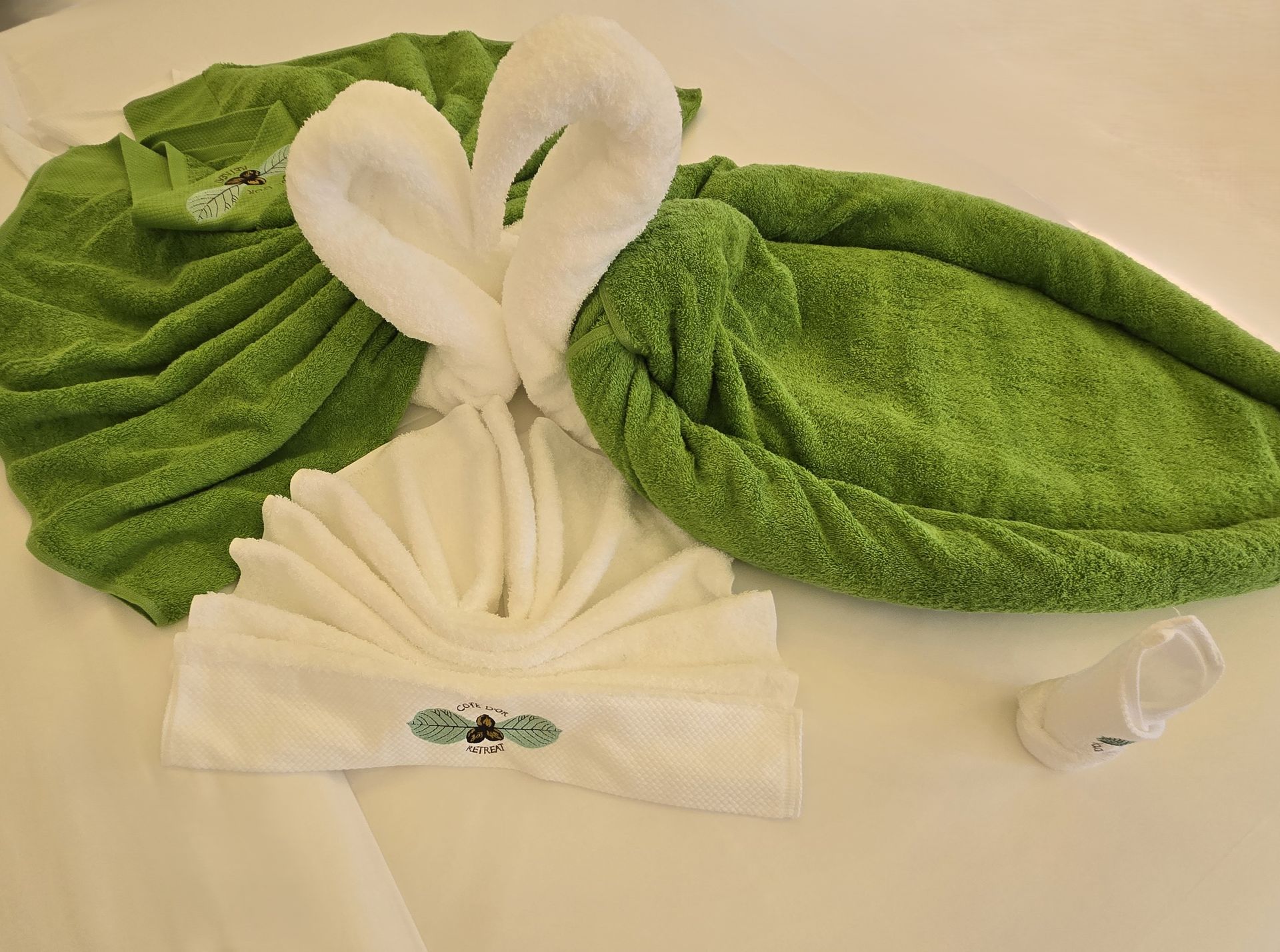 Green and white towel art on a bed: a swan, a leaf, and a head covering, plus a disposable cup.