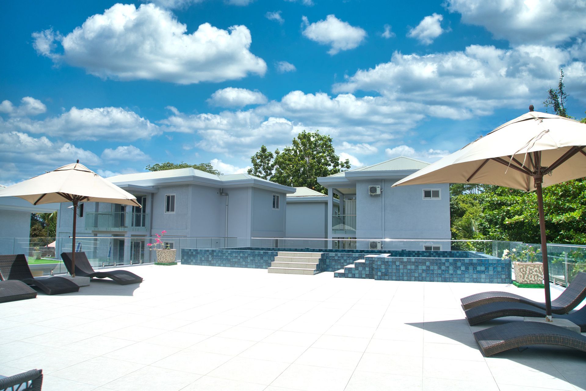 Poolside lounge area with umbrellas, chaise lounges, and light blue buildings under a cloudy sky.