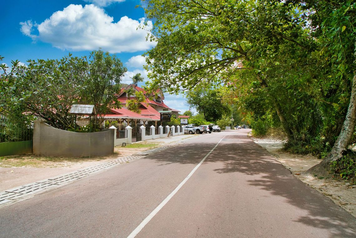 Paved road lined with trees, buildings with red roofs in the distance, blue sky with clouds.