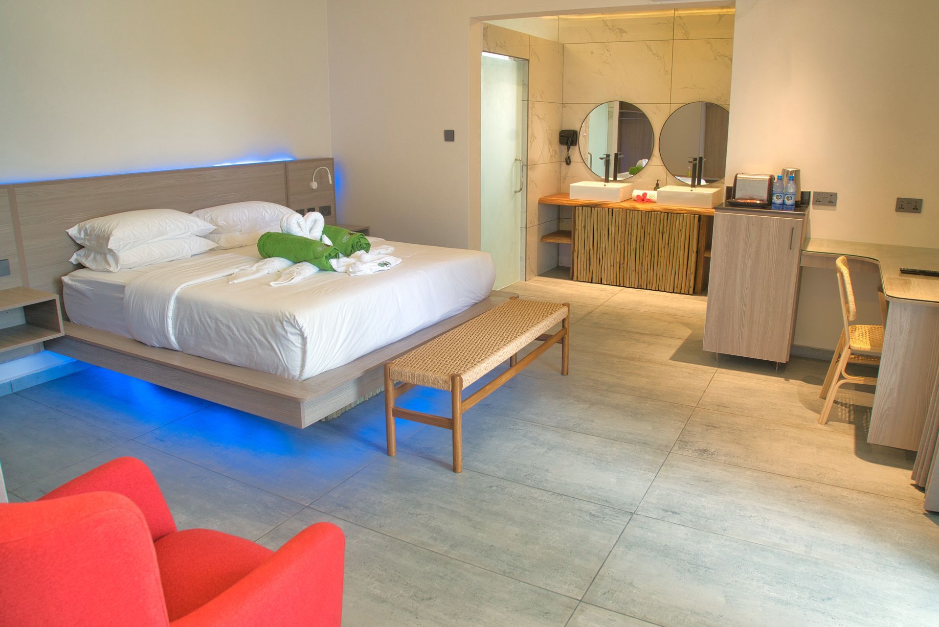 Modern hotel room with a bed, bench, and bathroom. Red chairs in foreground, blue lights under the bed.