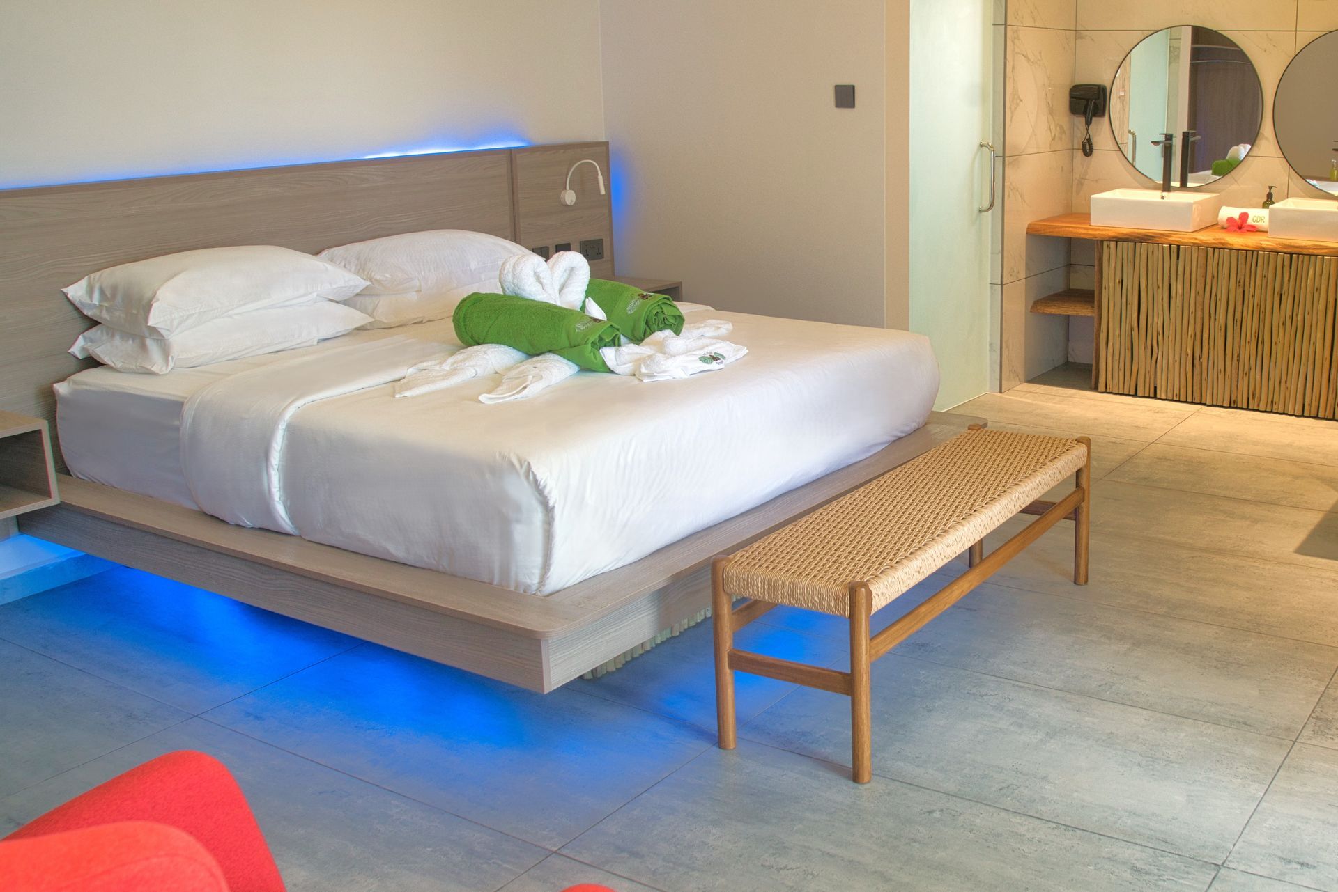 Bedroom with a bed, bench, and bathroom. Bed has white linens, blue lighting, and towels shaped like birds.