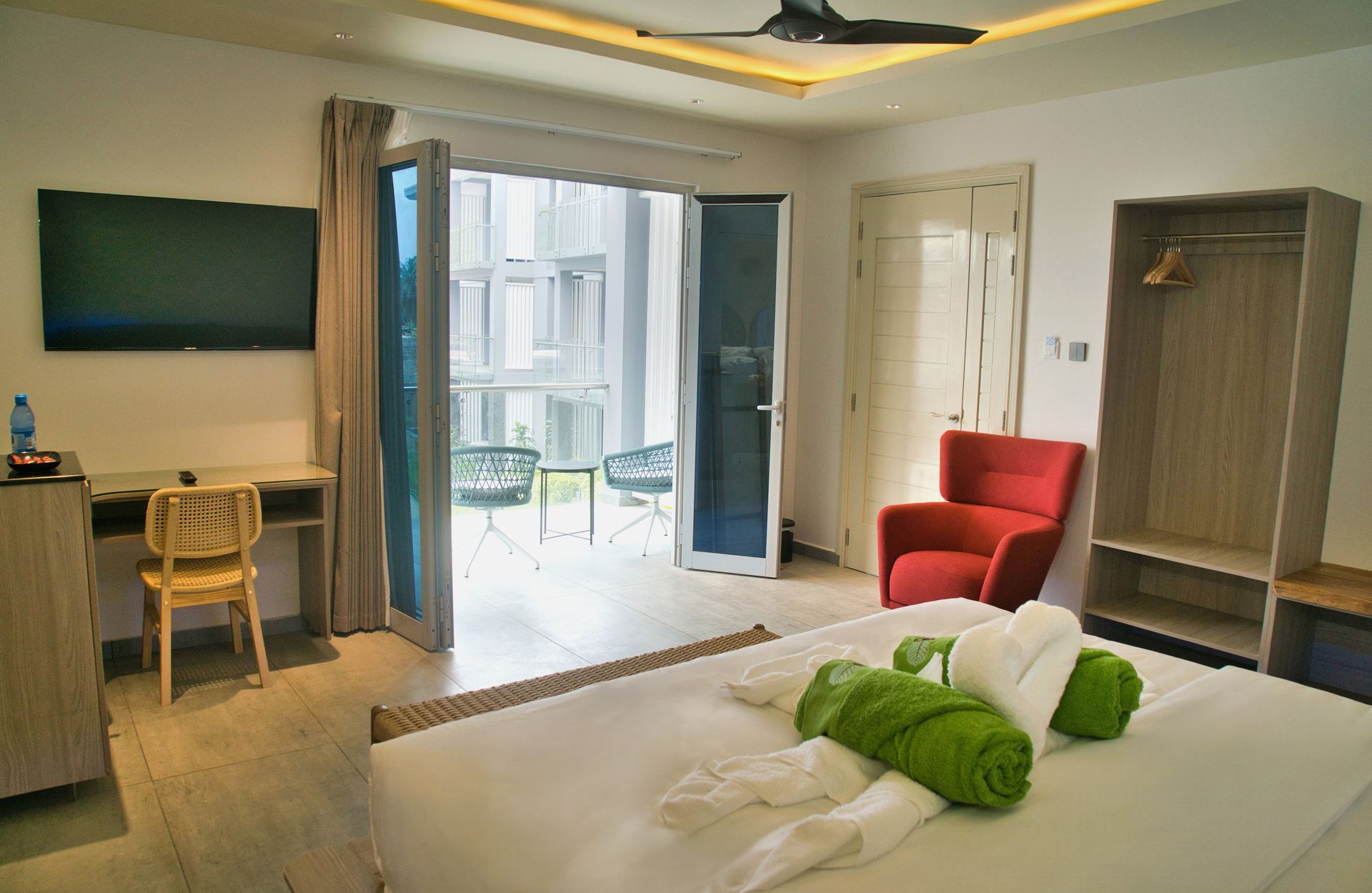Bedroom with balcony, bed with green towels, desk, TV, red armchair, closet.