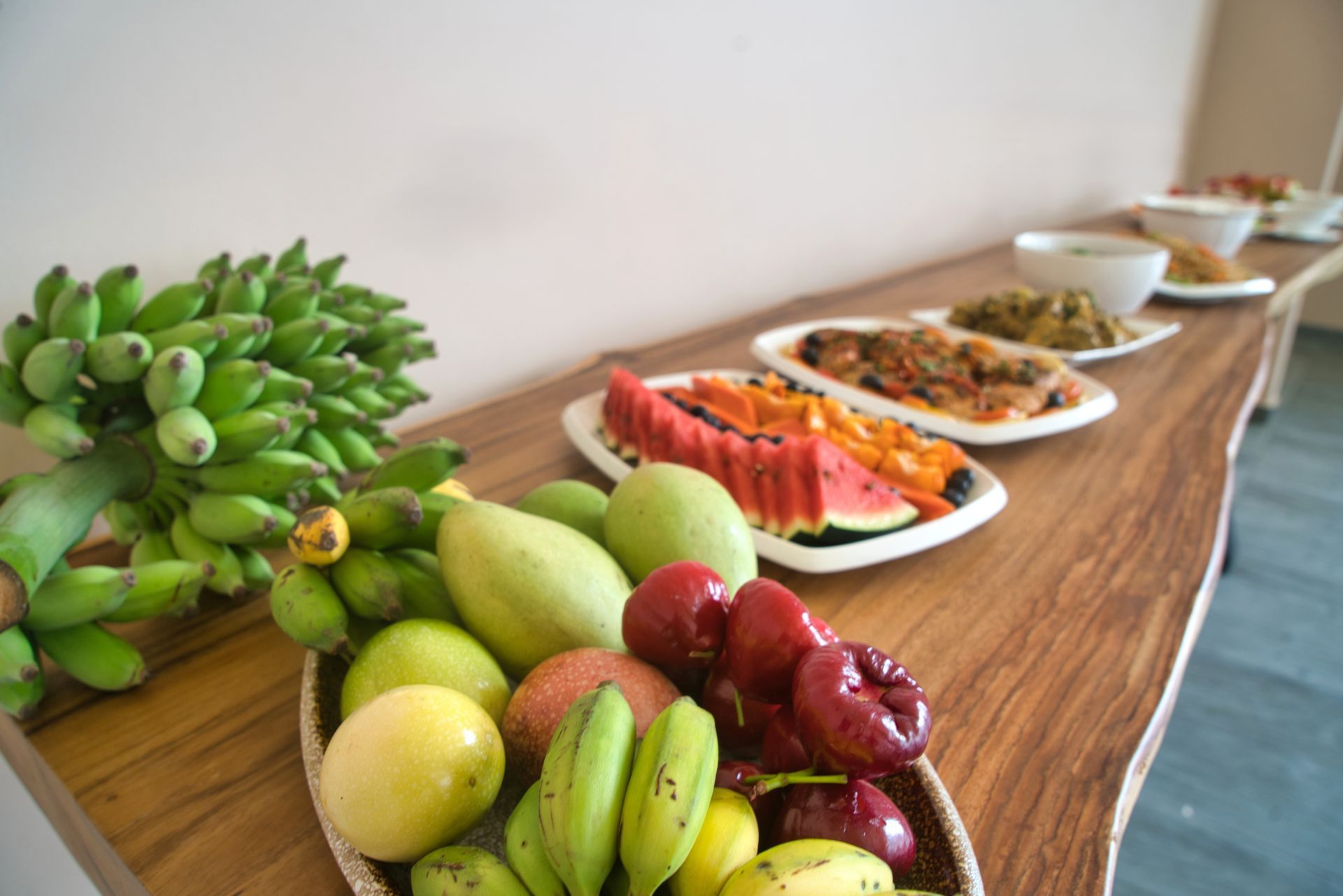 Fruits and dishes on a wooden buffet table: bananas, mangoes, watermelon, papaya, and cooked food.