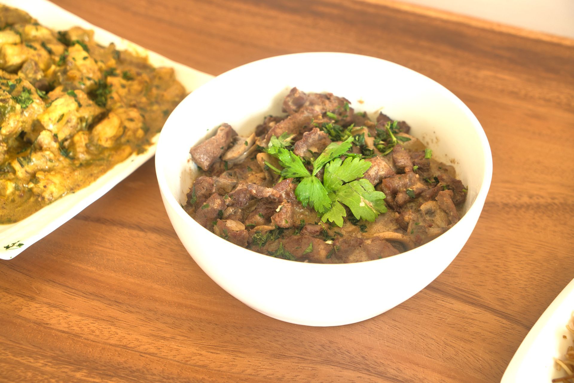Bowl of cooked meat topped with cilantro, alongside a dish of curry on a wooden table.