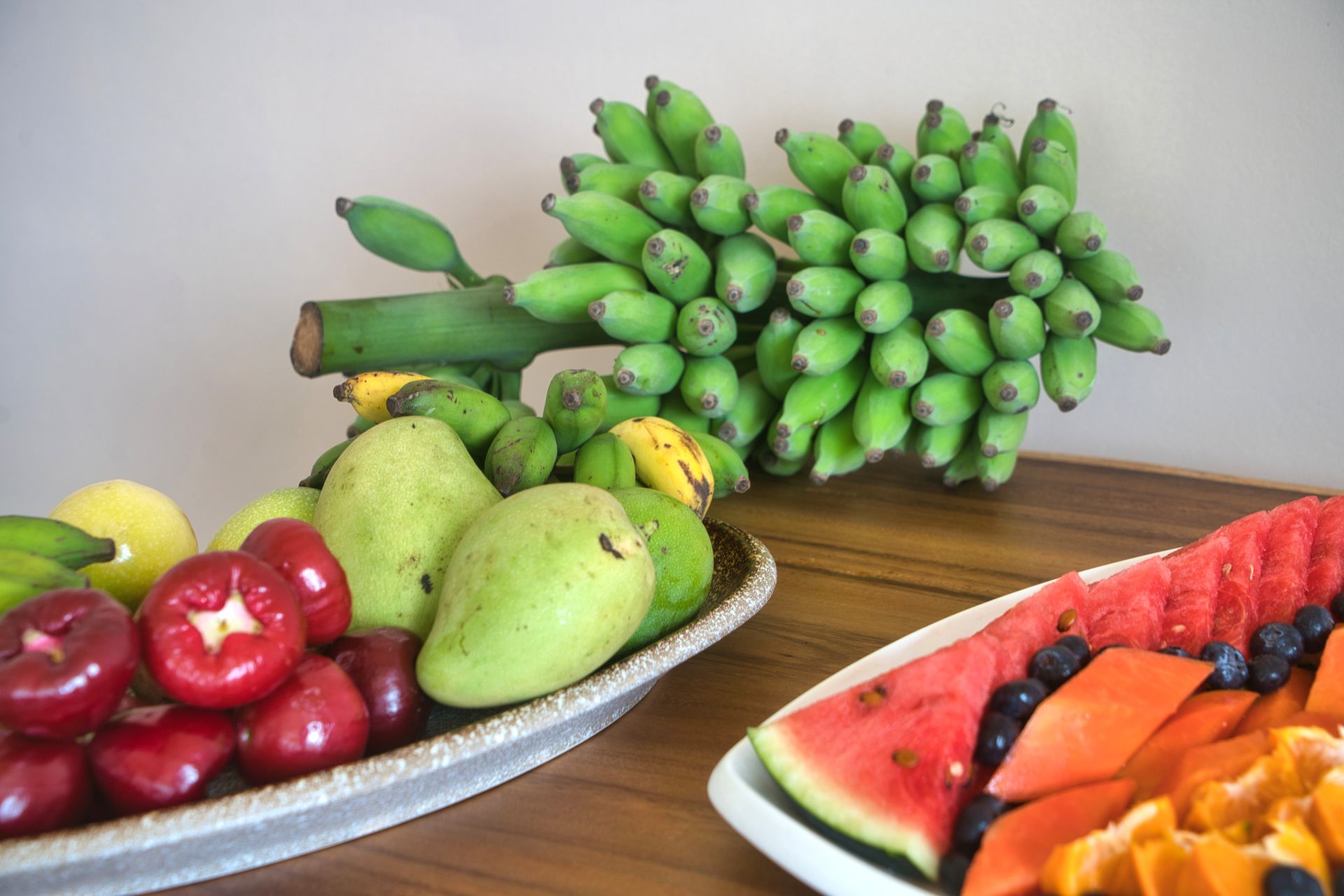 Platter of diverse tropical fruits: green bananas, mangos, watermelon, papaya, and more on a wooden table.