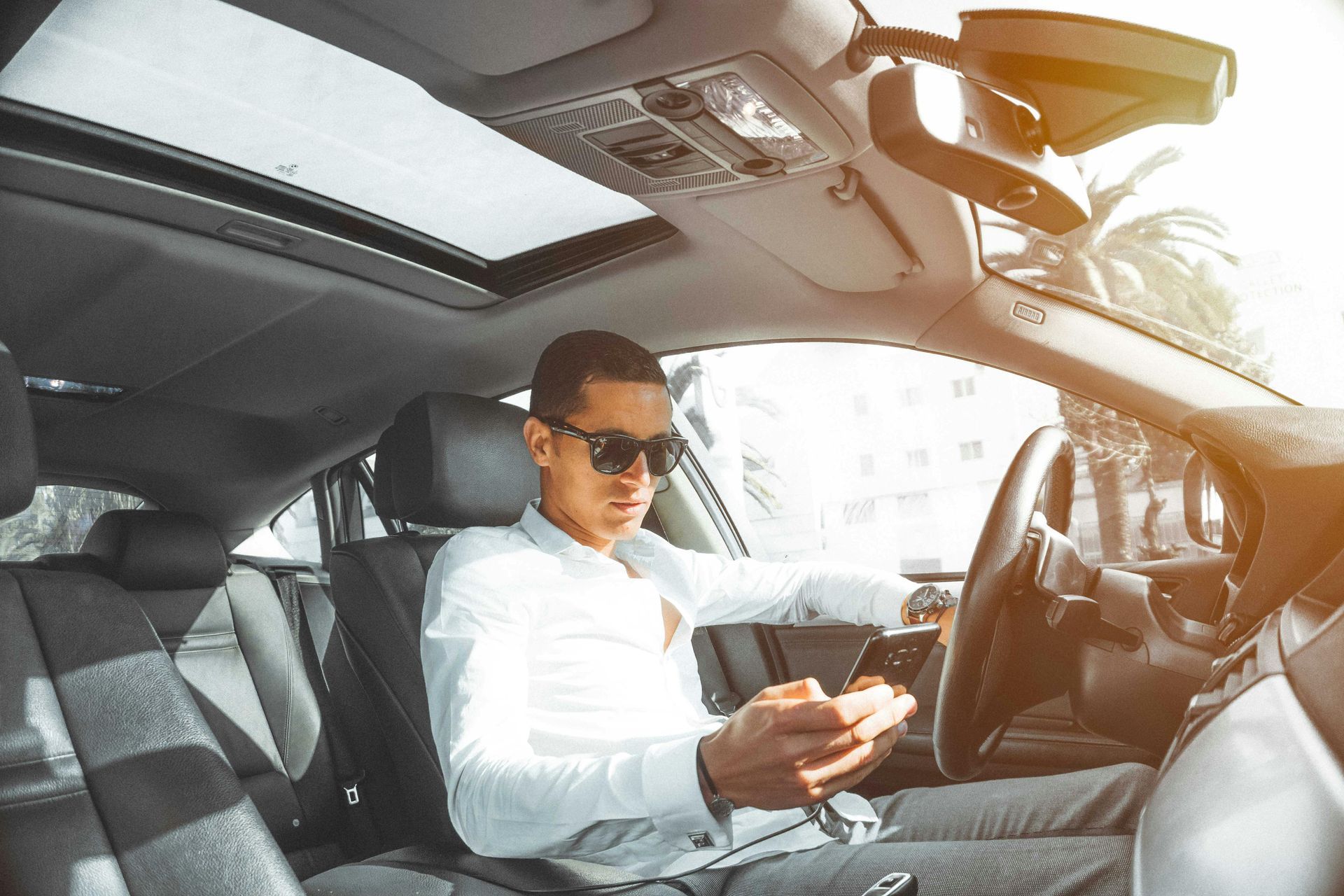 Man in sunglasses using phone while driving a car; sunlight streams through window.