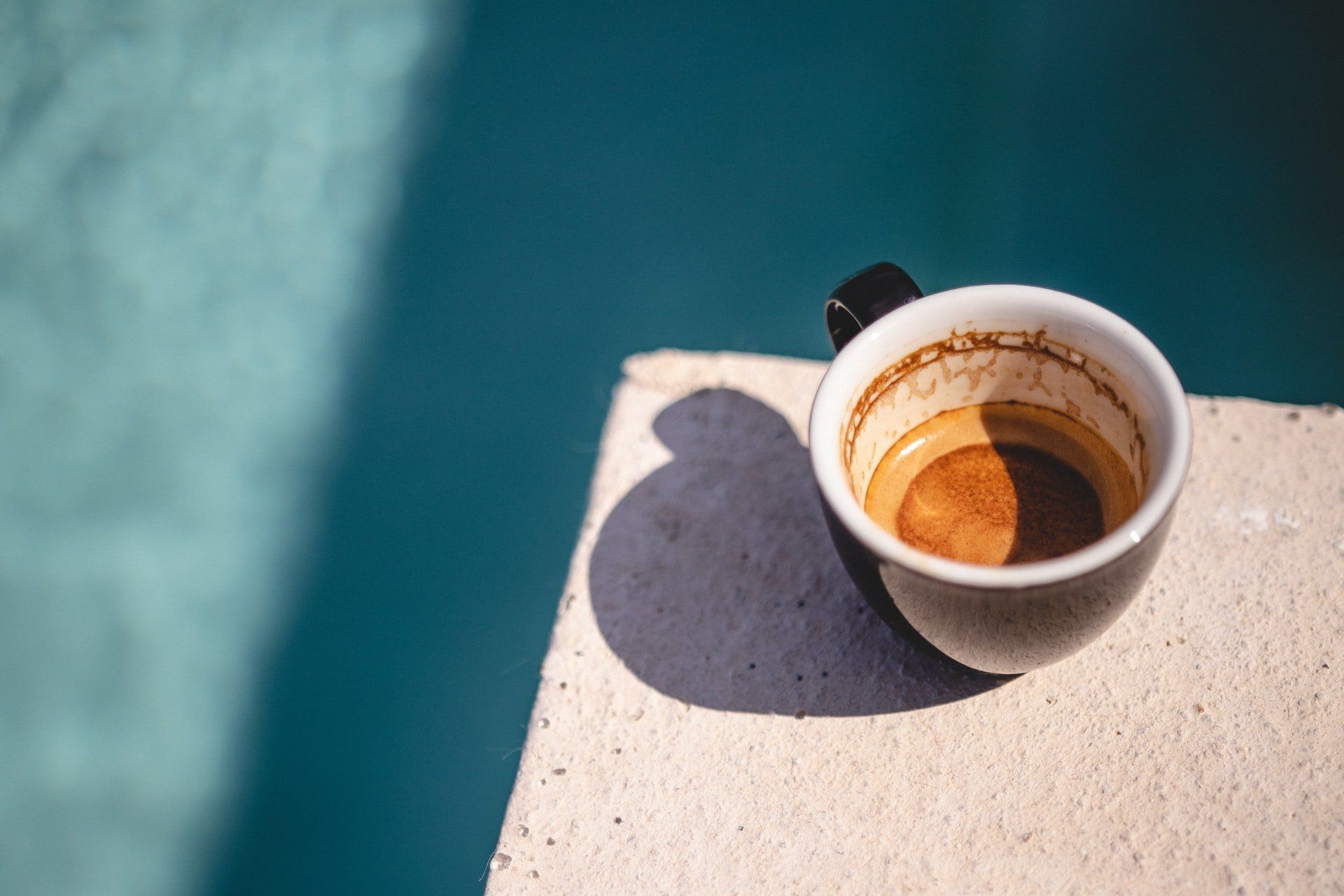 A mostly empty black coffee mug on a white ledge with a pool of blue water in the background.