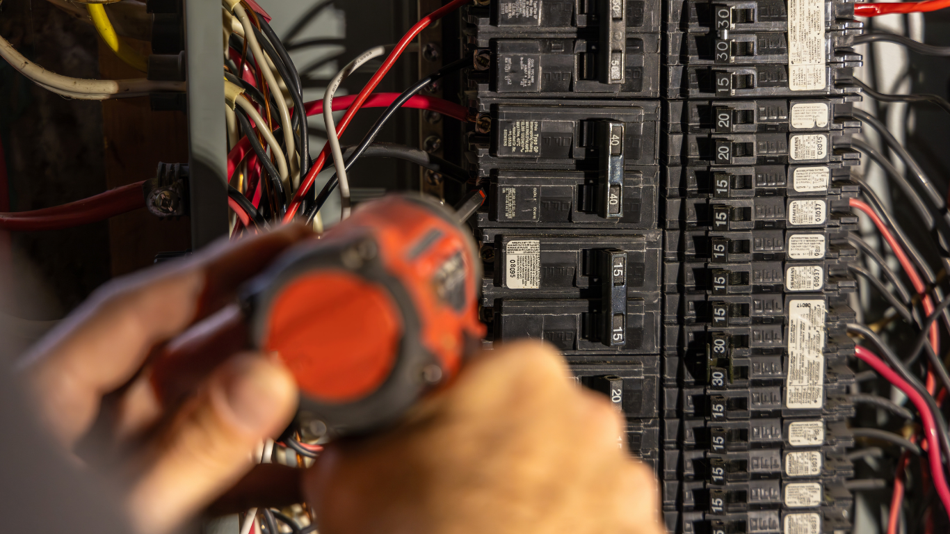 Hands using a power drill to tighten a screw on an electrical breaker panel.