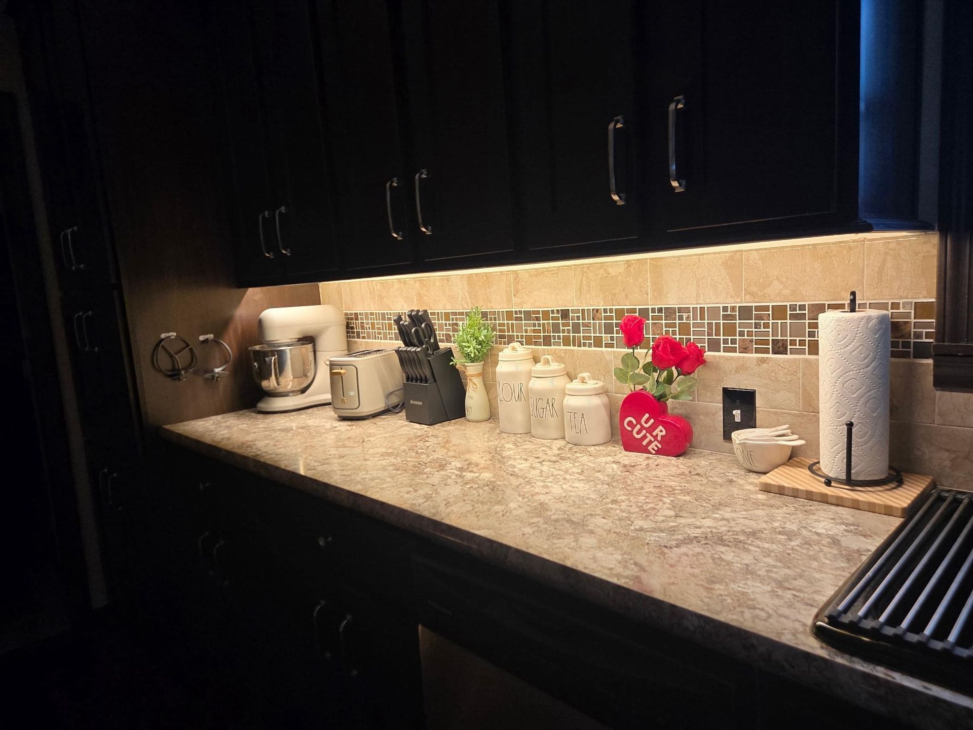 A kitchen counter with dark cabinets, granite countertops, under-cabinet lighting, appliances, and decorative items.