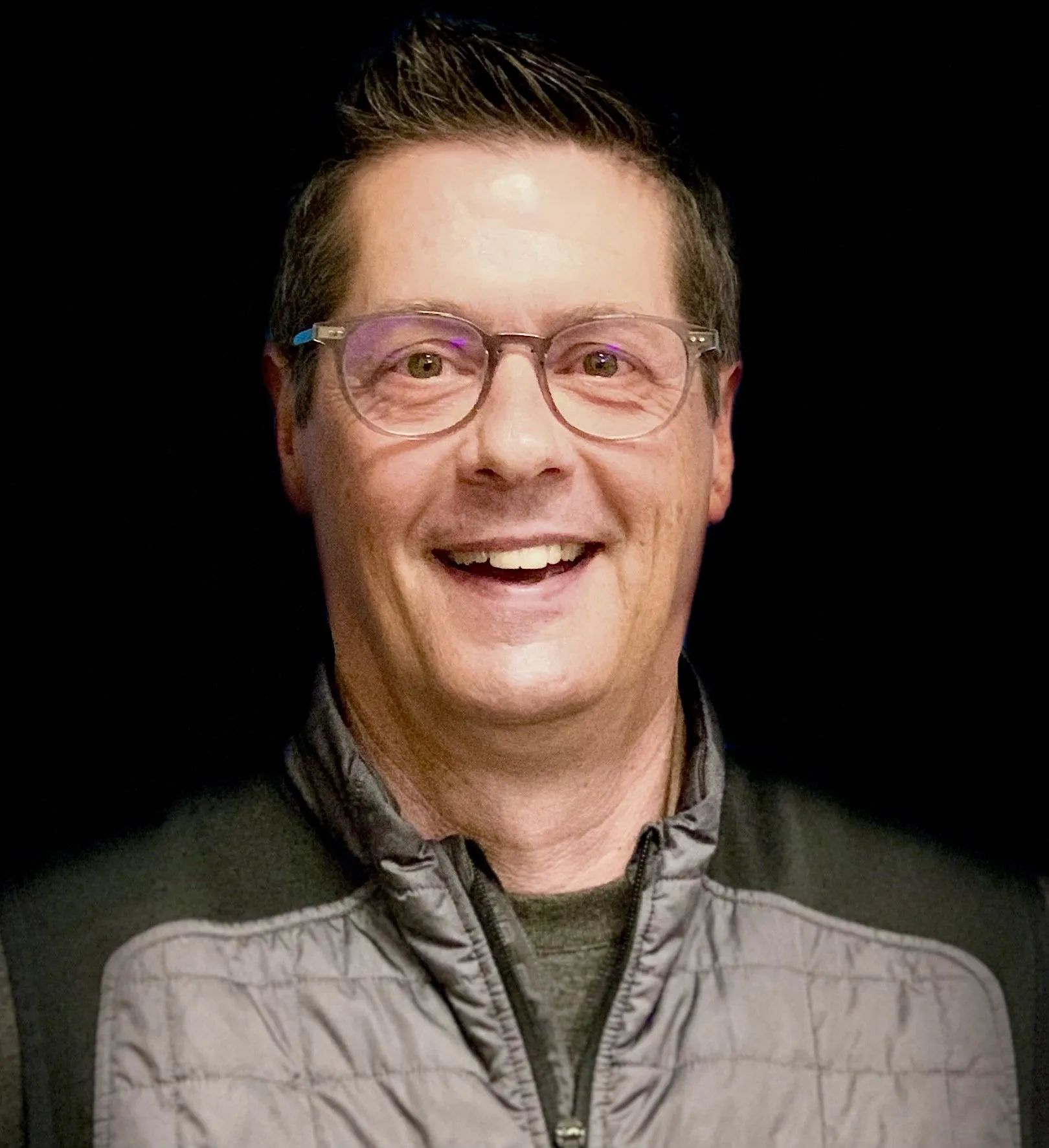 A man wearing glasses and a jacket is smiling for the camera.