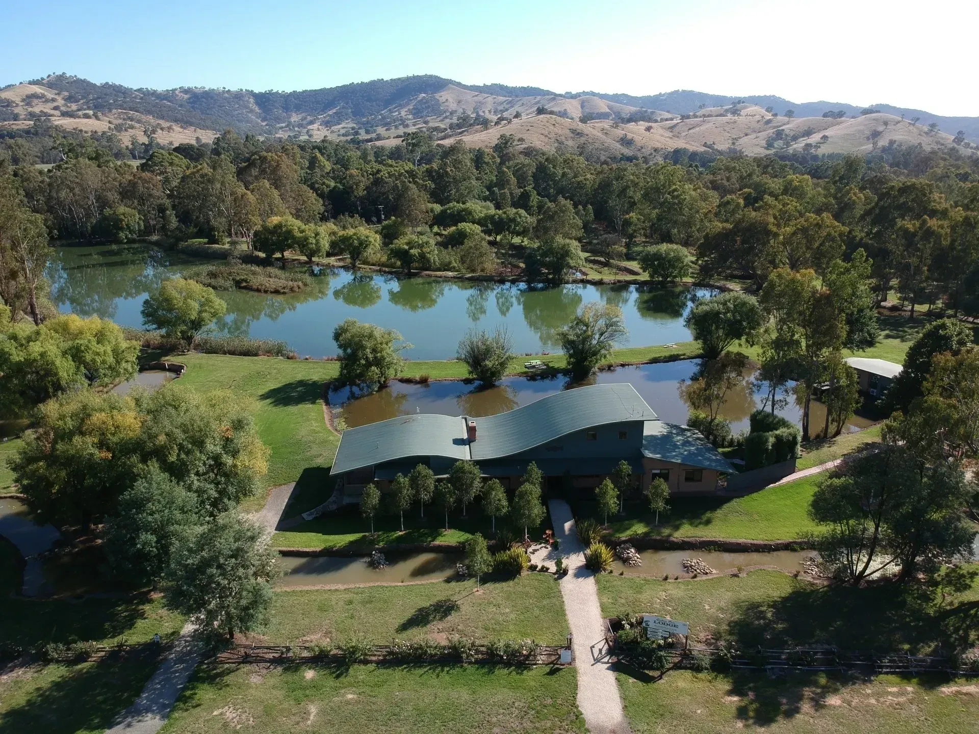 A house with a dark, wavy roof sits beside a pond, surrounded by lush trees and rolling hills under a clear blue sky.
