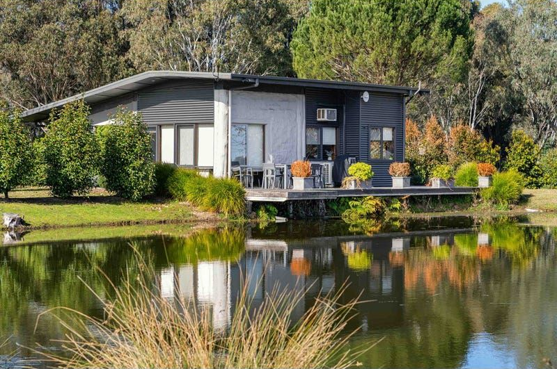A modern house with dark siding and a deck overlooking a calm pond surrounded by trees and vegetation.