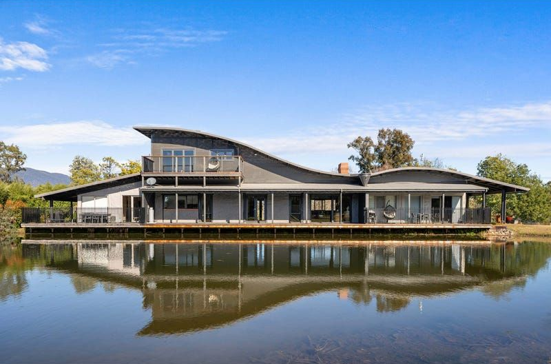 A modern house with a unique curved, wave-like roof sits beside a calm, reflective pond under a clear blue sky.