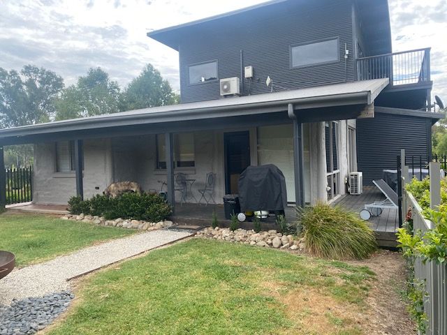 A modern, grey two-story house with a wooden deck overlooking a calm lake surrounded by lush green trees.