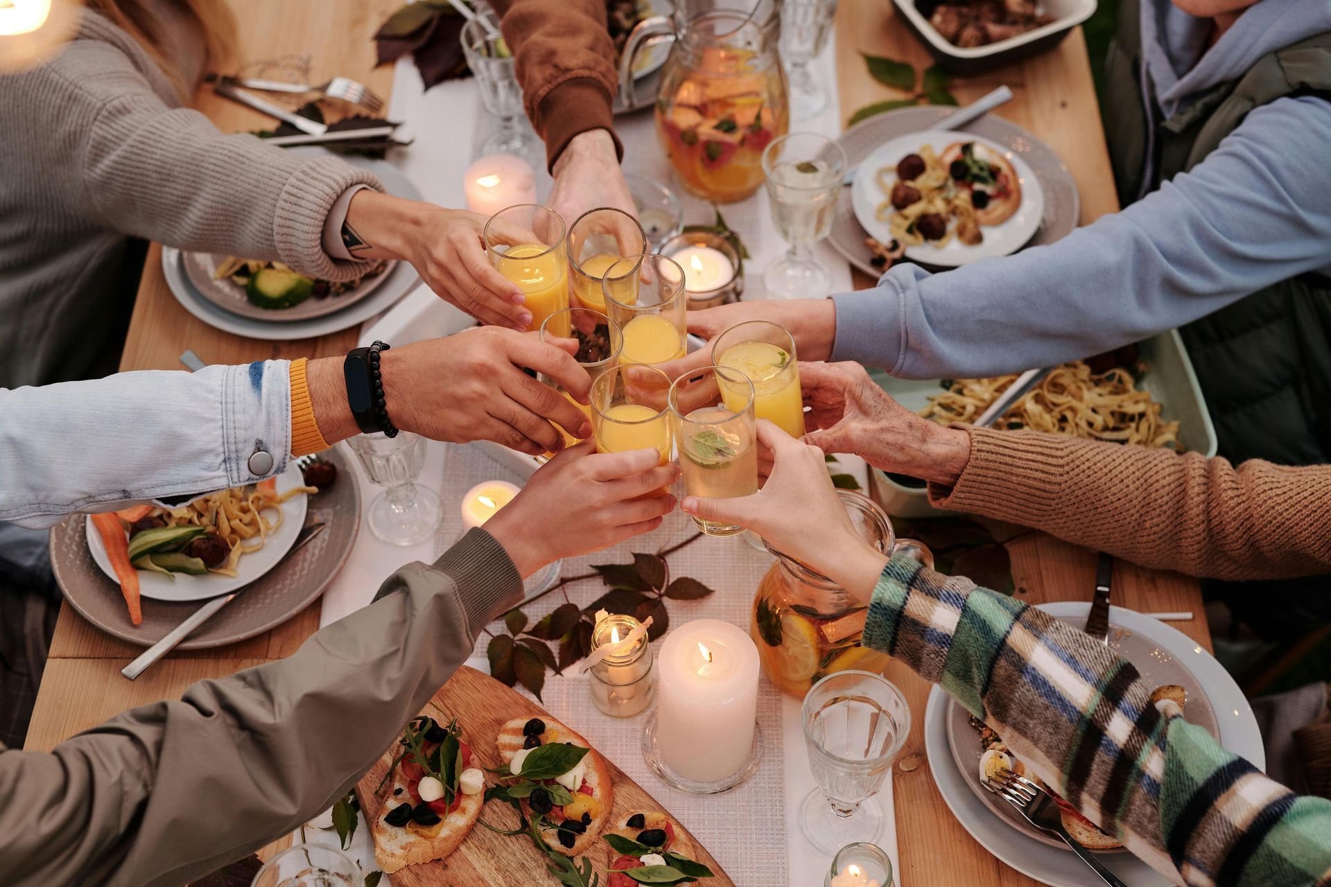 People gathered around a wooden table, toasting with glasses of orange-colored drinks, with food and candles nearby.