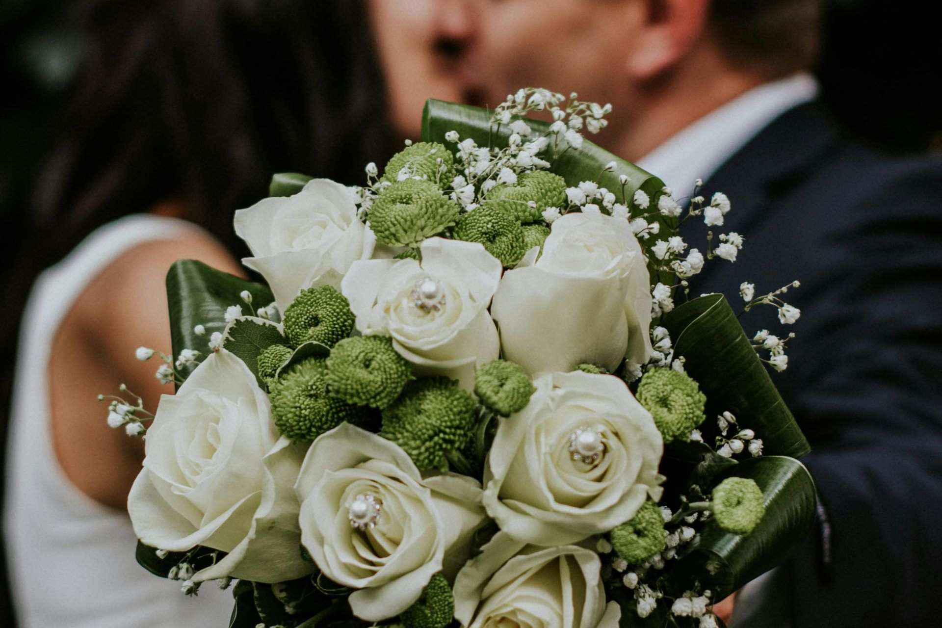 A close-up of a wedding bouquet featuring white roses with embedded crystals, green filler flowers, and baby's breath.
