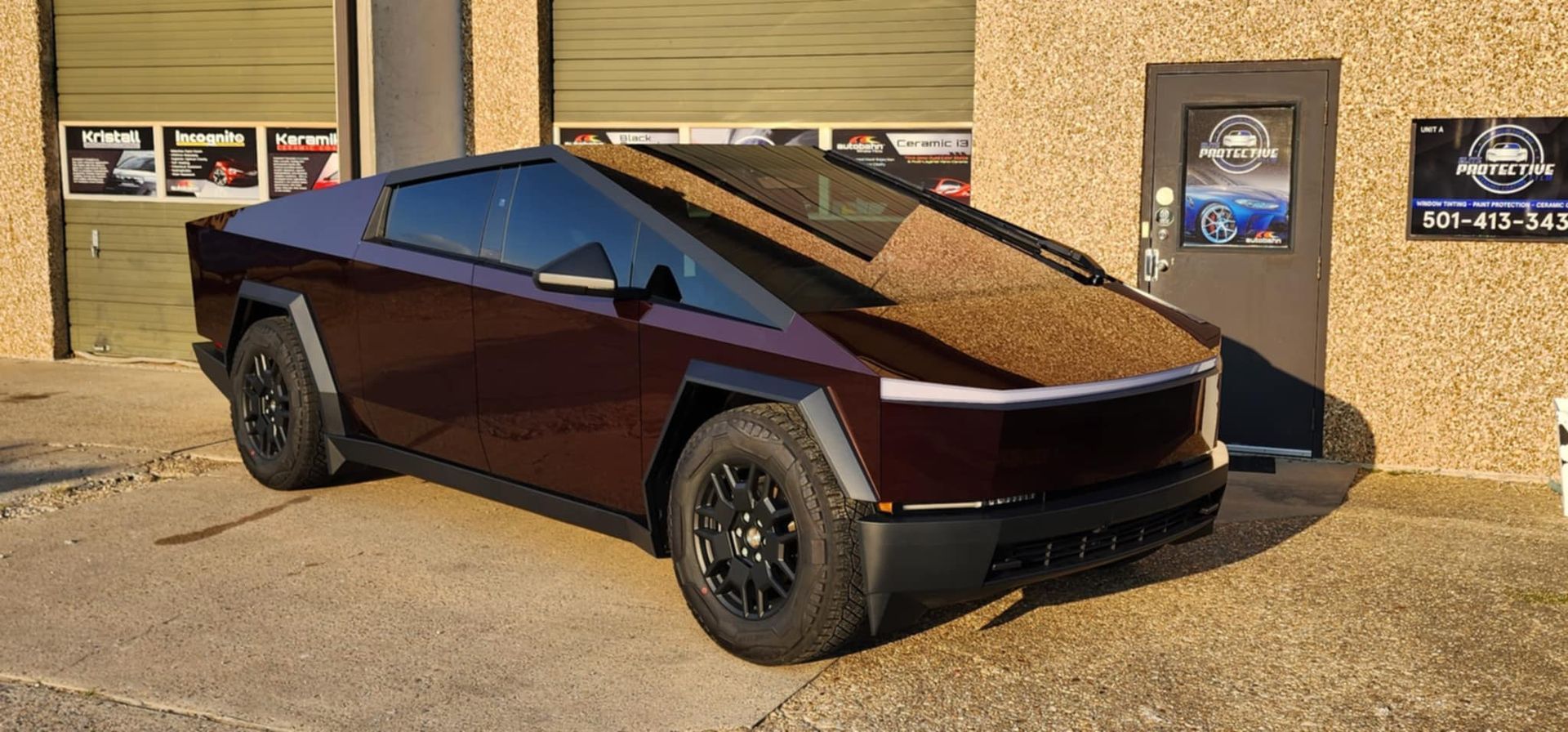 A dark red Tesla Cybertruck parked in front of a tan brick building with garage doors.