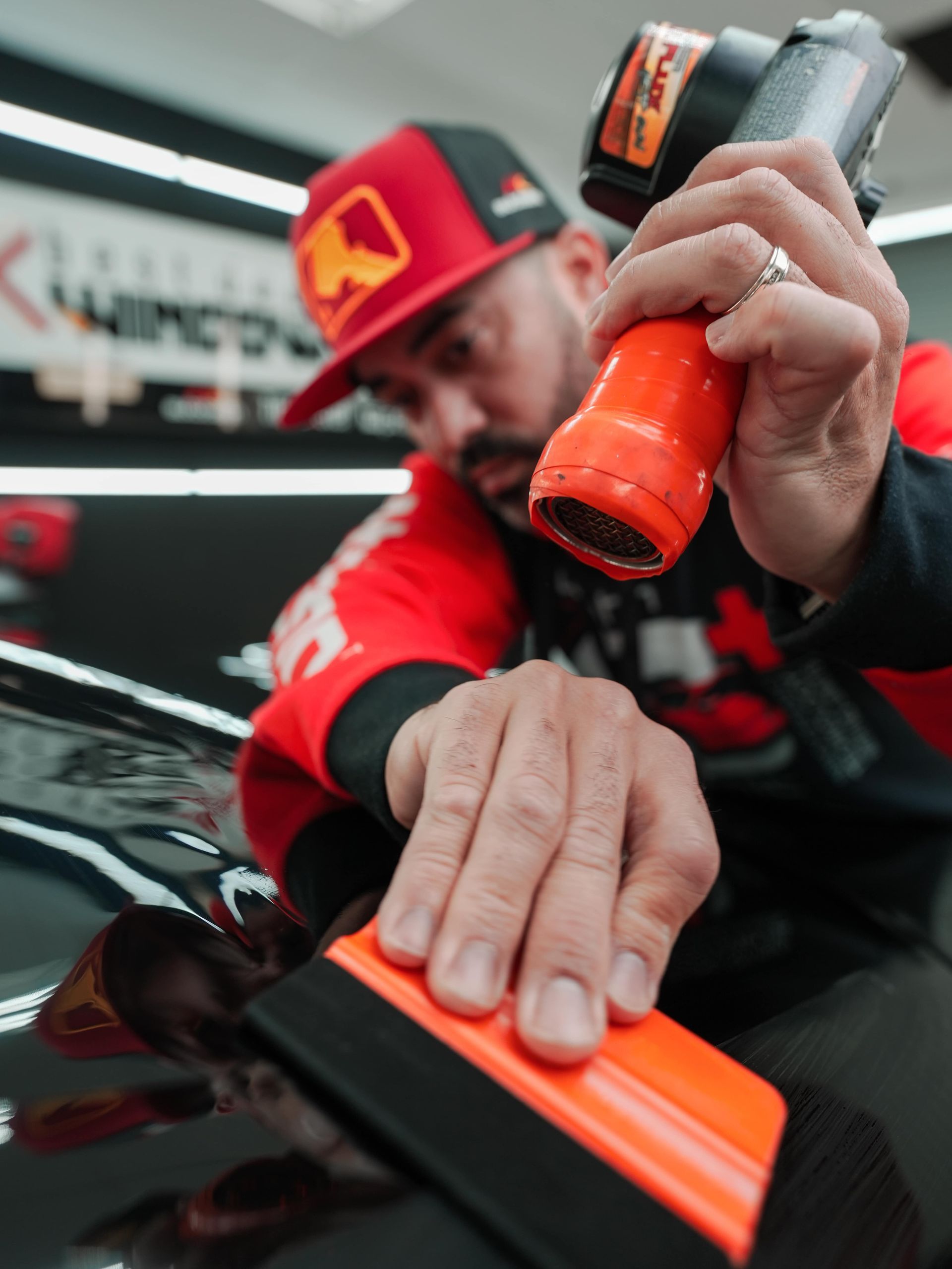 Man in red cap using a heat gun and squeegee to apply vinyl wrap on a black car.