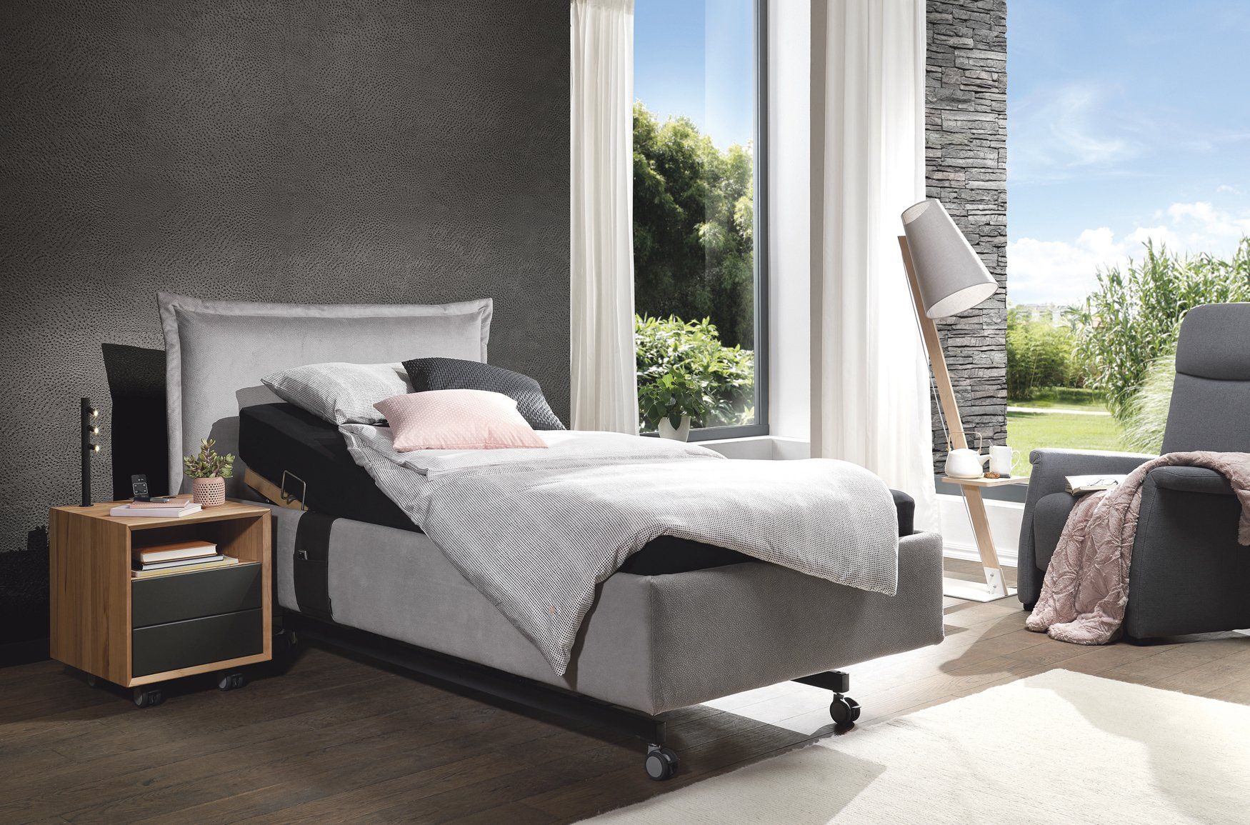 Schlafzimmer mit verstellbarem Bett, Nachttisch und Sessel neben einem großen Fenster. Neutrale Farben, natürliches Licht.