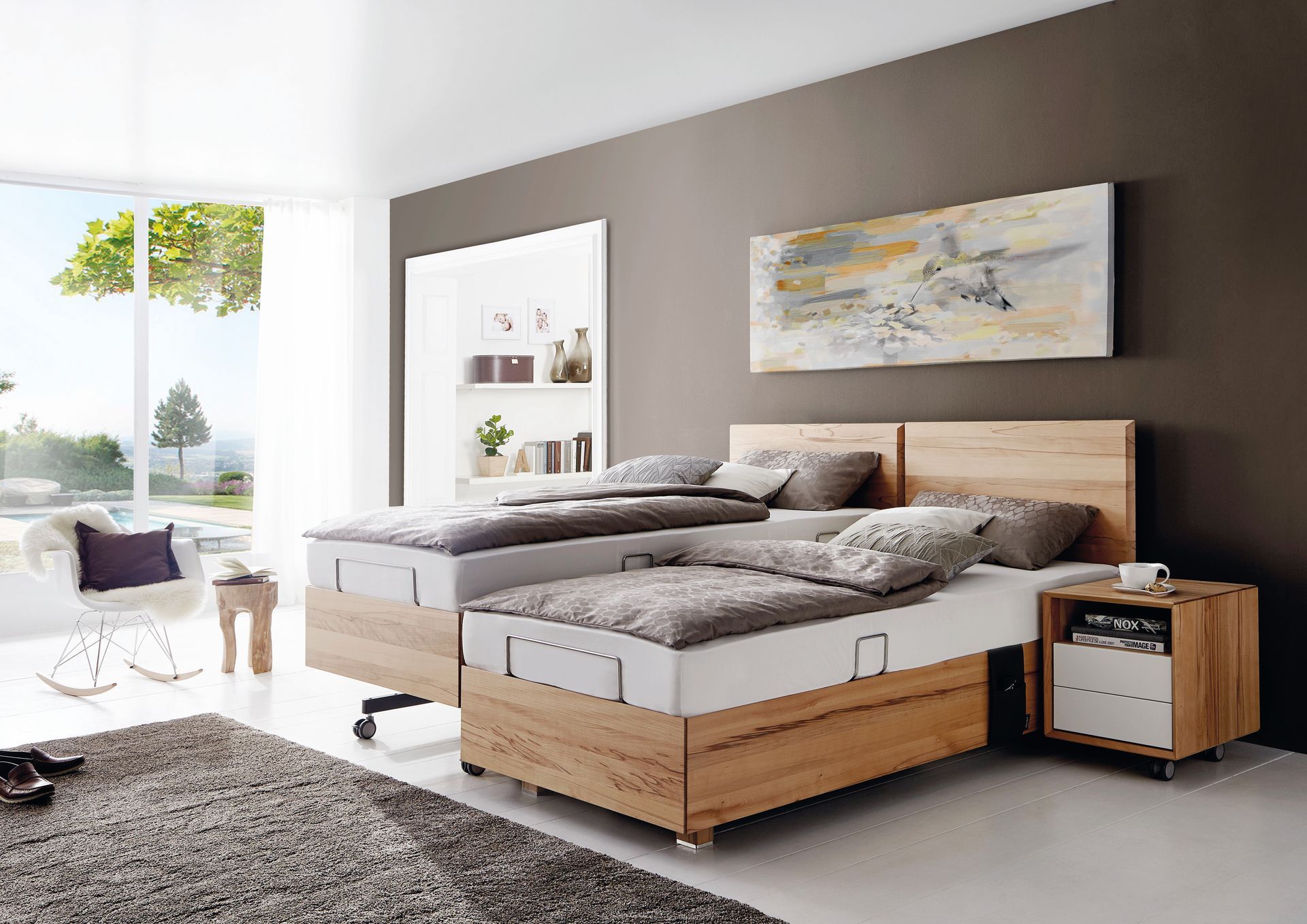 Modernes Schlafzimmer mit zwei Betten, einem Nachttisch, Kunstwerken und einem großen Fenster mit Blick auf einen Hinterhof.