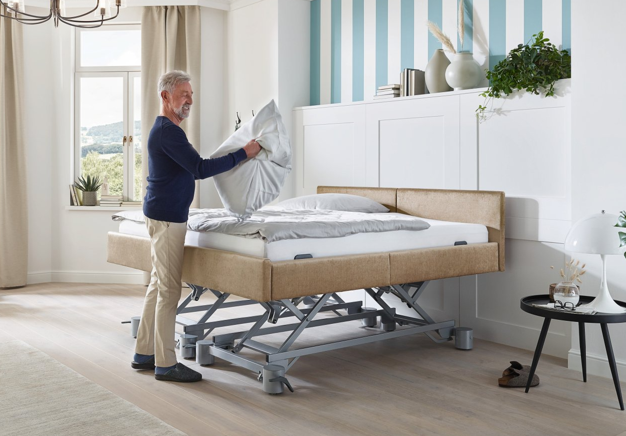 Ein Mann in einem blauen Pullover legt ein Kissen auf ein beiges verstellbares Bett in einem Schlafzimmer.