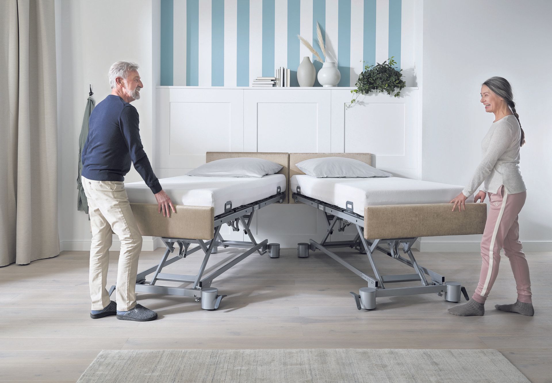 Ein älterer Mann und eine ältere Frau heben zwei verstellbare Betten in einem hellen Schlafzimmer mit gestreiften Wänden an.