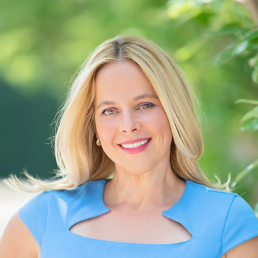 Smiling blonde woman in a light blue dress outdoors with green foliage background