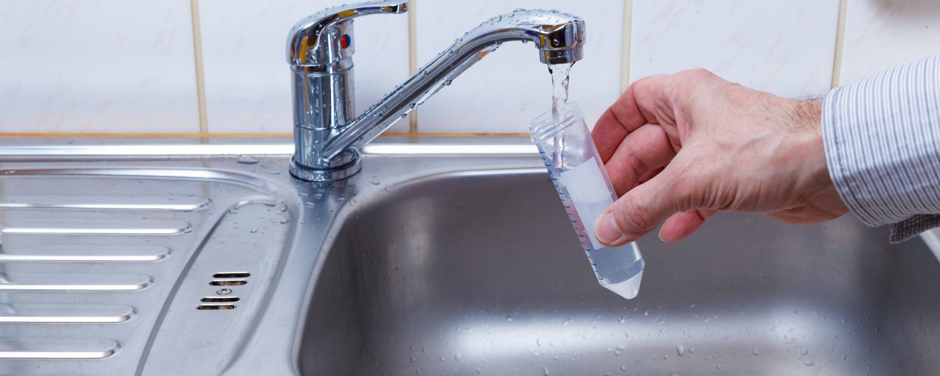 Une main tenant un flacon transparent sous l'eau courante dans un évier en acier inoxydable.