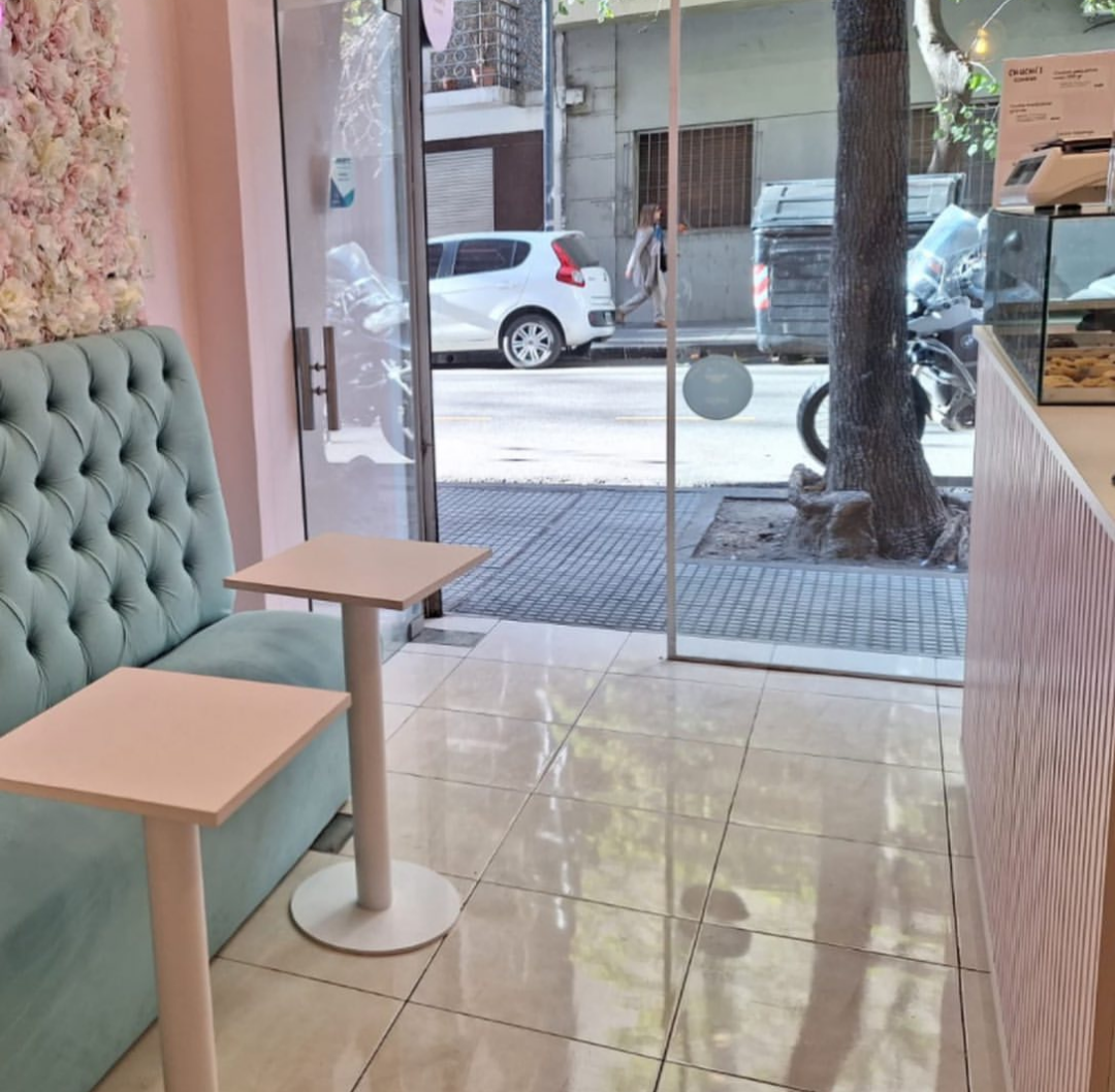 Interior de una cafetería rosa y verde azulado con mesas, un banco acolchado y una puerta de vidrio abierta a la calle.