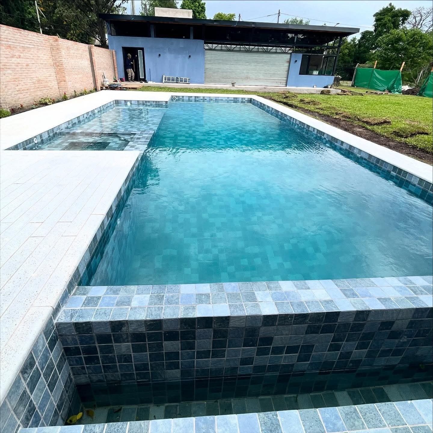 Piscina en el patio trasero con agua azul cristalina, borde de azulejos y un pequeño edificio al fondo.