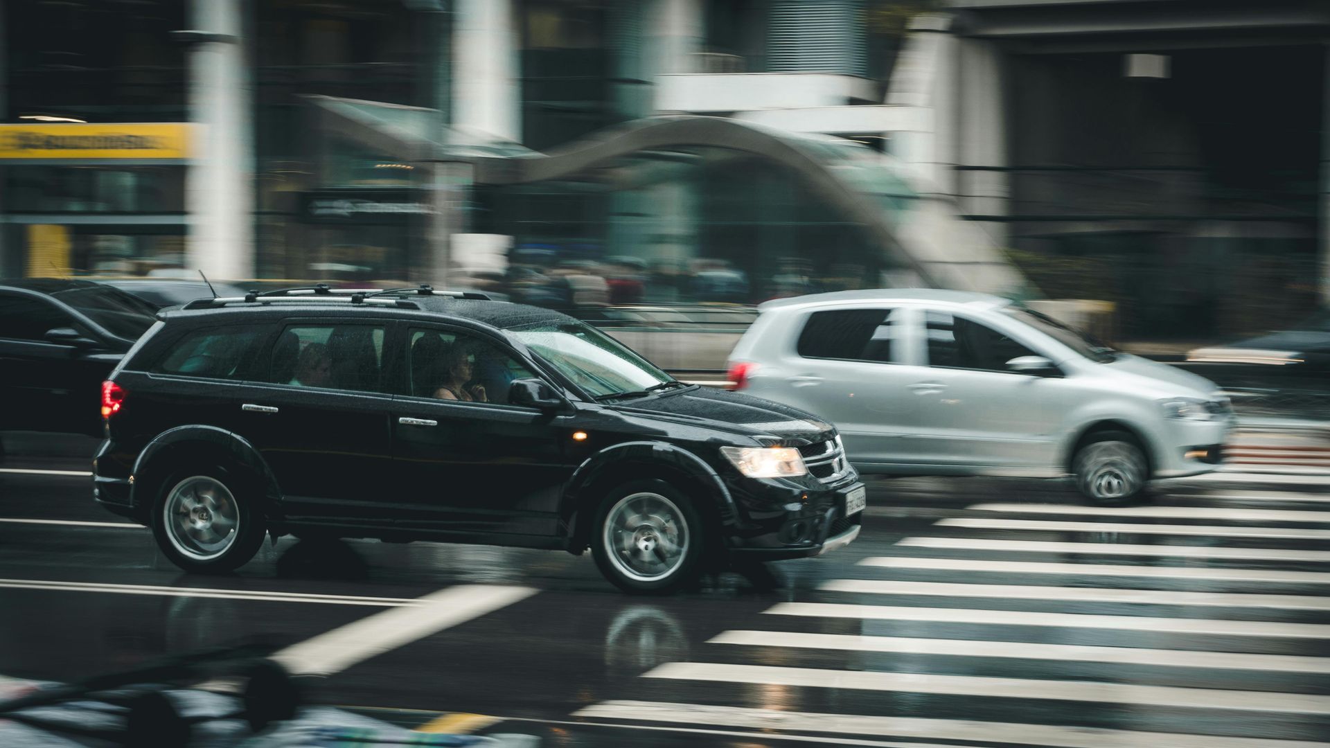 A black suv and a white van are driving down a wet city street.